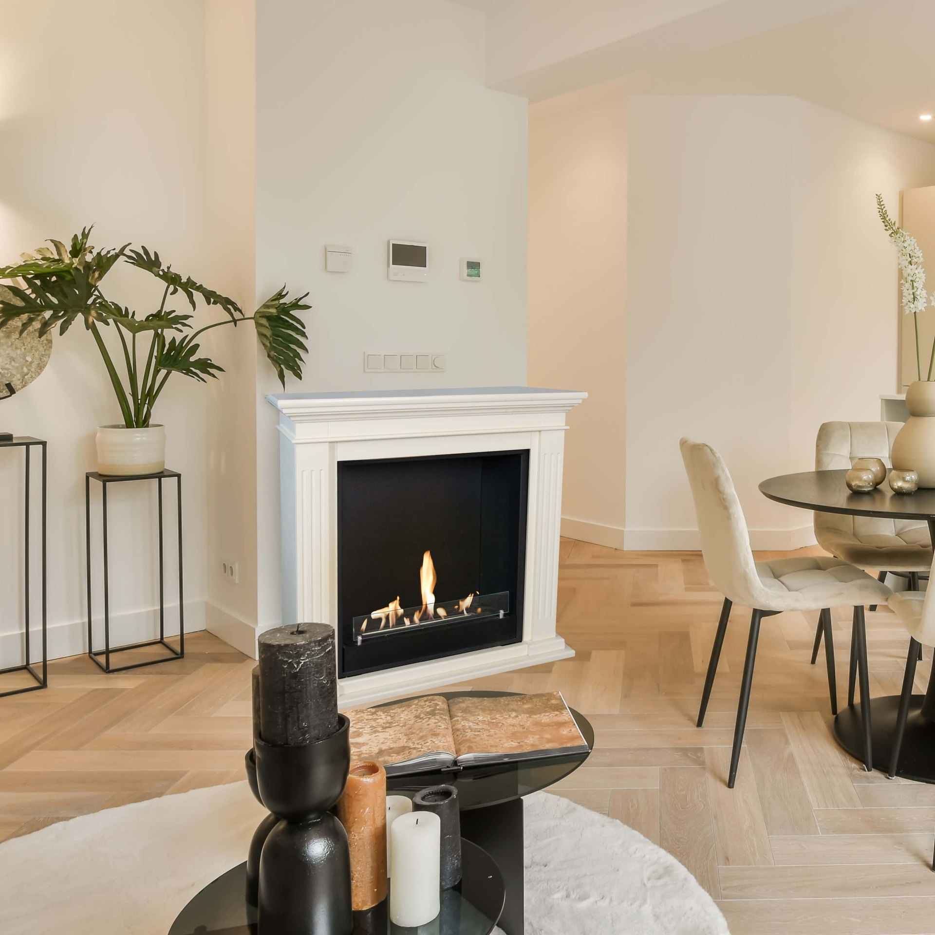 Een woonkamer met een open haard, kaarsen en een tafel. Links een plant, rechts een eettafel.