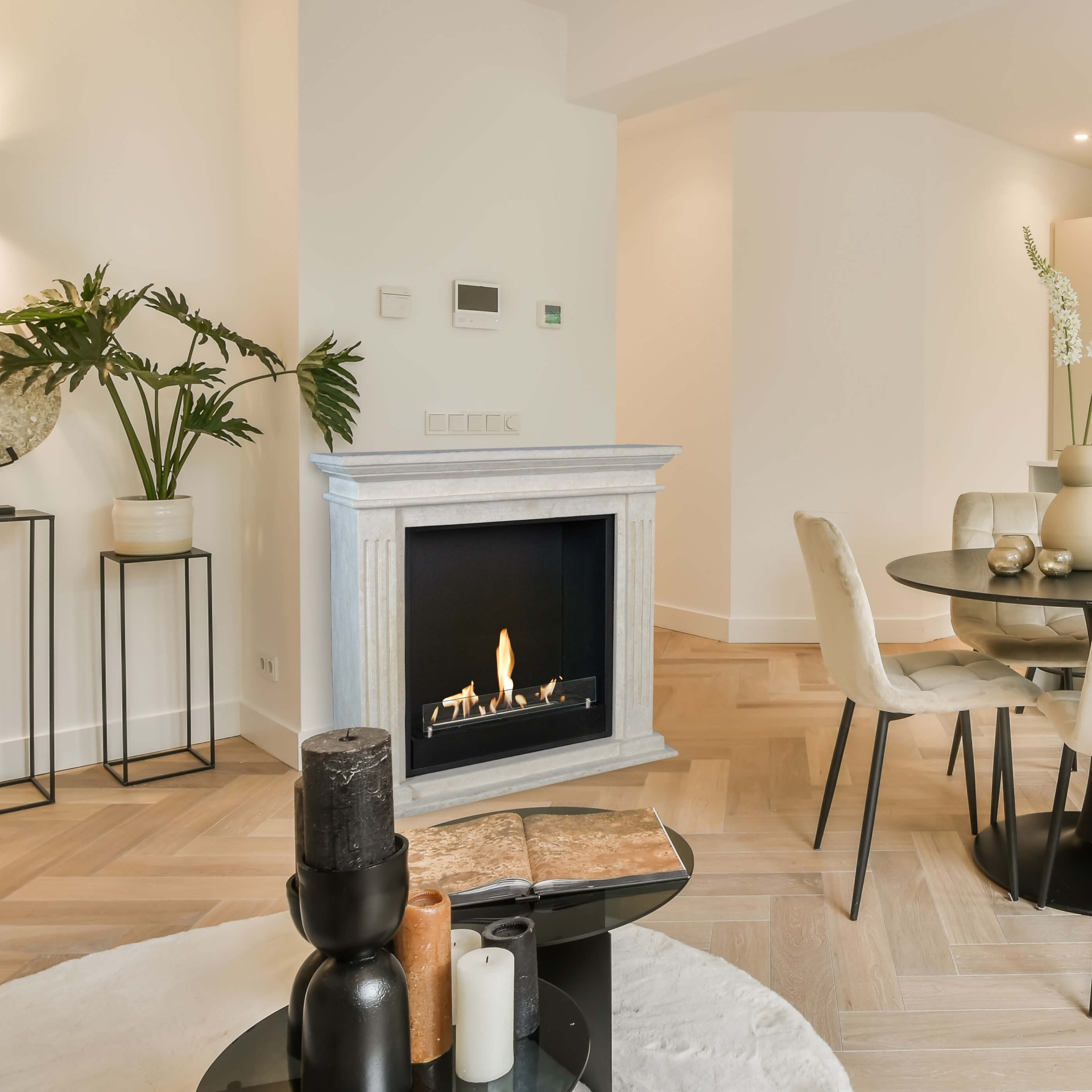 Binnenaanzicht van een woonkamer met open haard, tafel, stoelen, planten en kaarsen. Warme kleuren en natuurlijk licht.