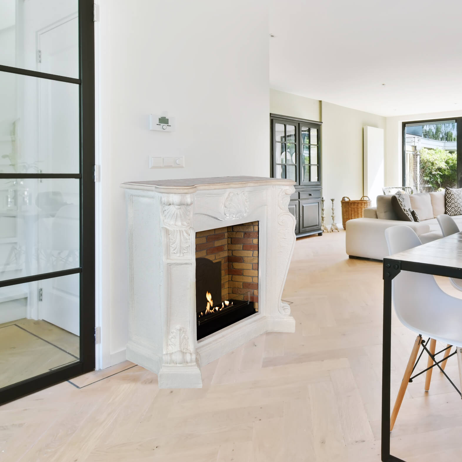 Een woonkamer met een witte open haard, een deur met zwart frame en een eettafel met witte stoelen. Een bank op de achtergrond.