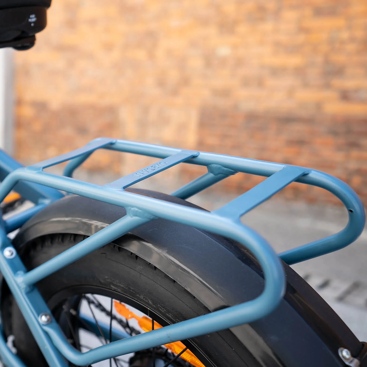 Widok z tyłu niebieskiego bagażnika na rowerze z czarną oponą i ceglanym tłem.
