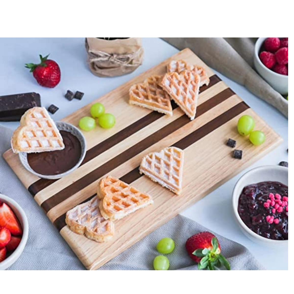 Herzförmige Waffeln mit Beeren, Schokolade und Trauben auf einem Holzbrett. Hintergrund hell.