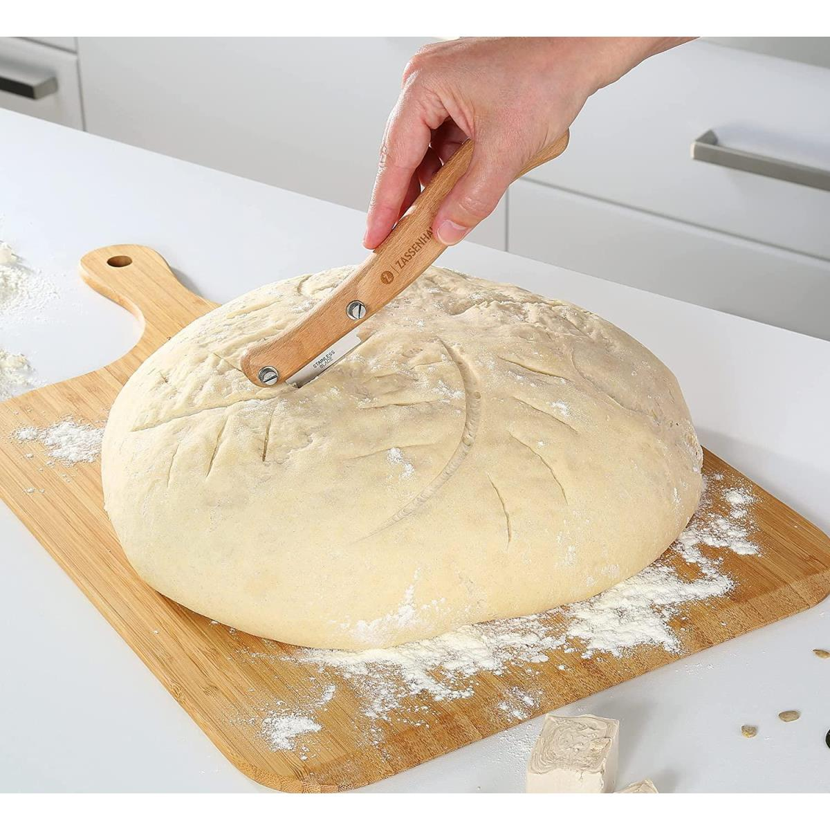 Eine Hand, die einen Brotlaib auf einem Holzschneidebrett mit einem Spezialmesser einschneidet.
