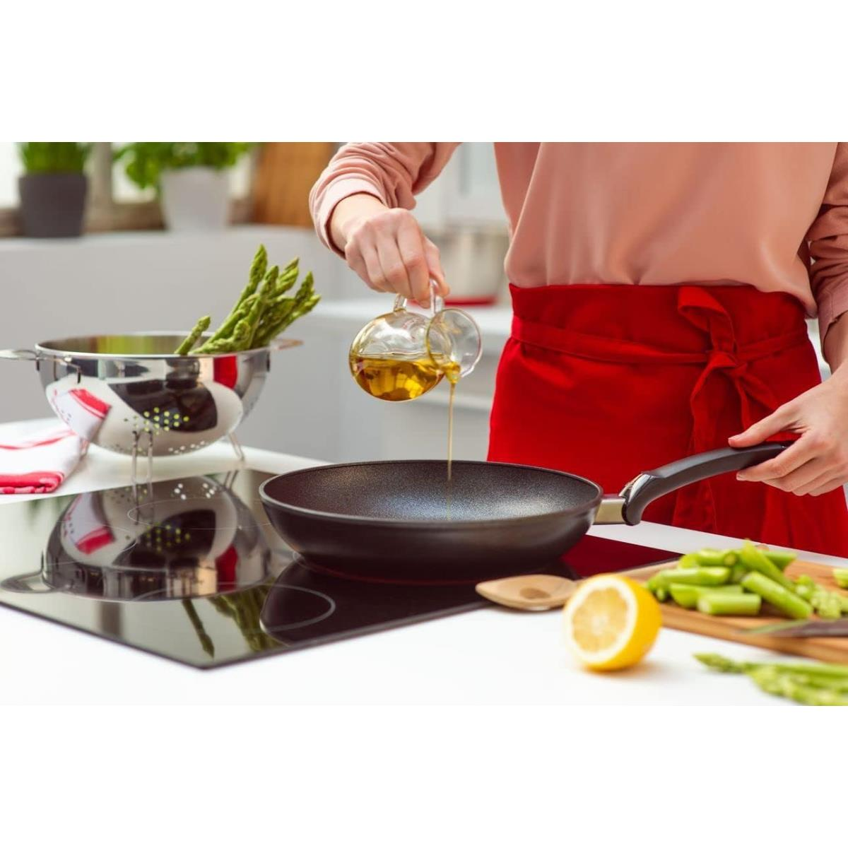 Persona che versa olio da una caraffa di vetro in una padella nera. Asparagi e limone presenti.