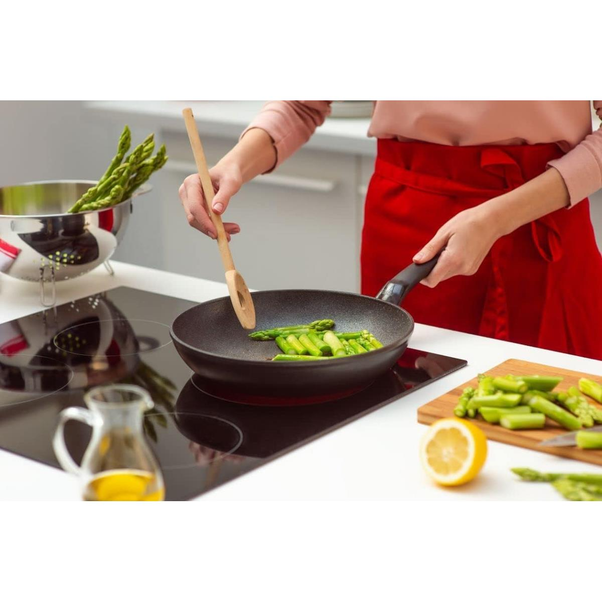Donna che cucina asparagi in una padella su un piano cottura nero, con olio e limone nelle vicinanze.
