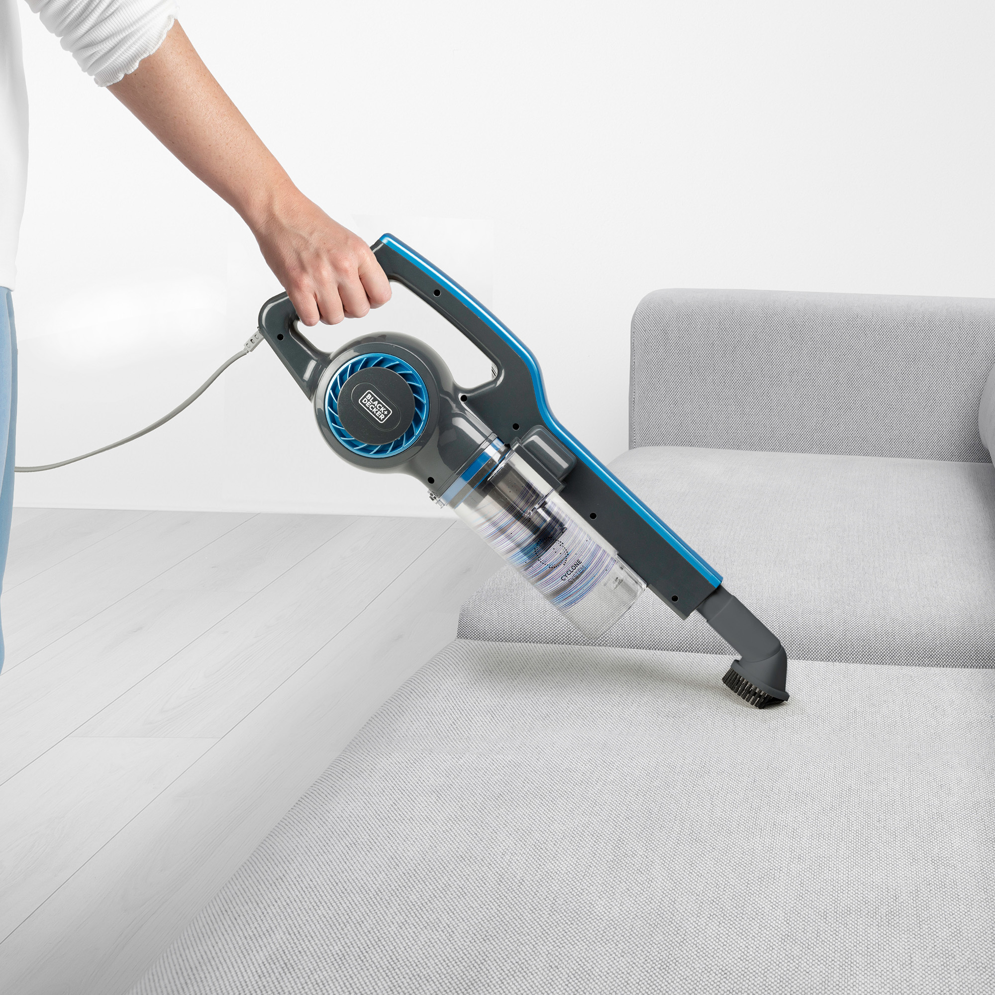 Ręka używająca odkurzacza ręcznego na szarej sofie, biała podłoga w tle.