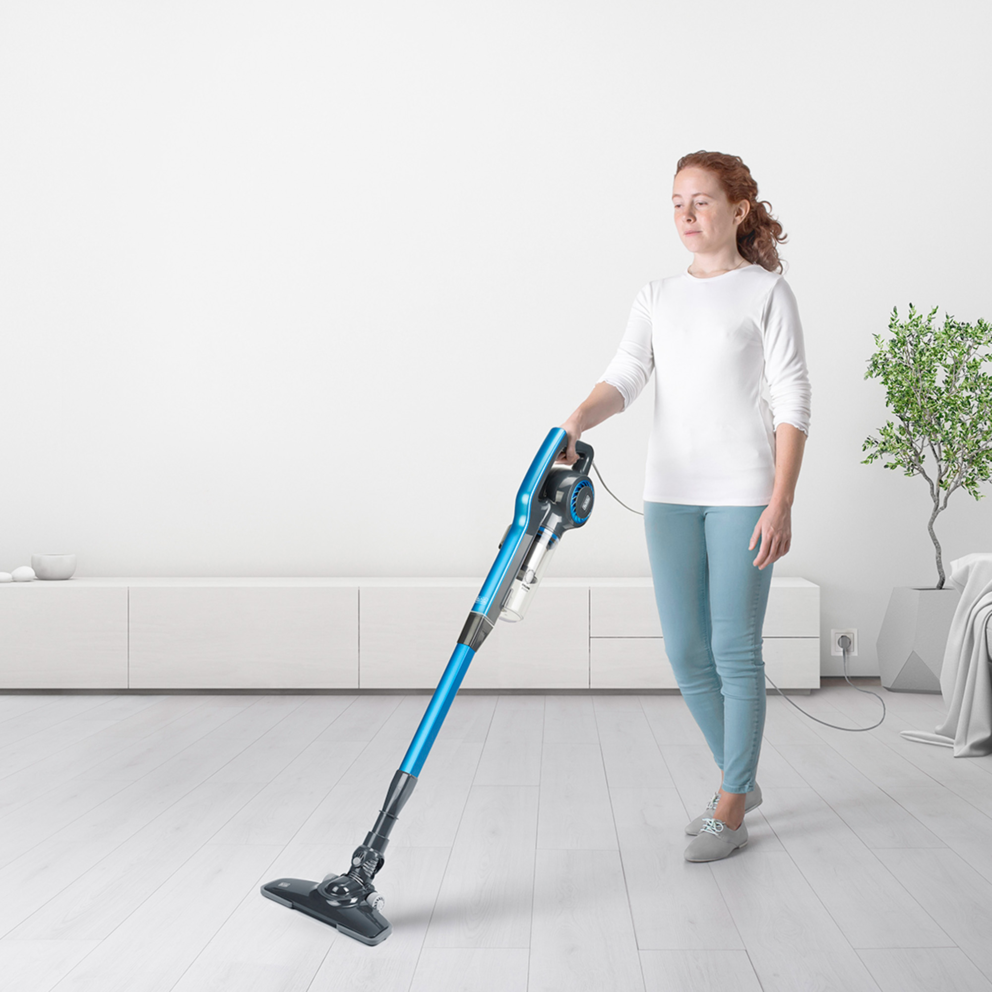 Kobieta czyści podłogę niebieskim odkurzaczem. Tło: biała ściana, roślina i meble.