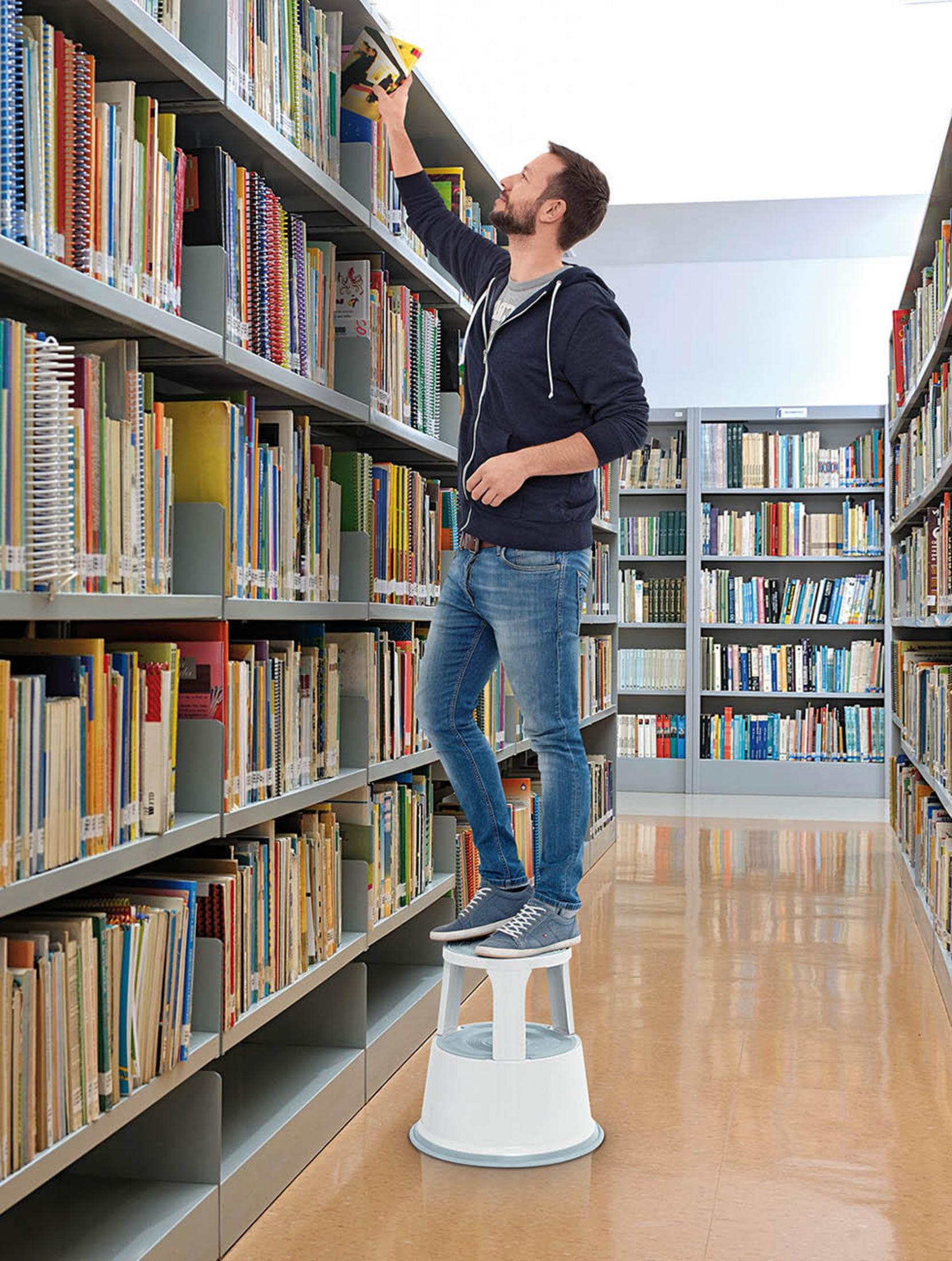 Ein Mann auf einem Tritthocker in einer Bibliothek, der nach einem Buch in einem Regal greift.