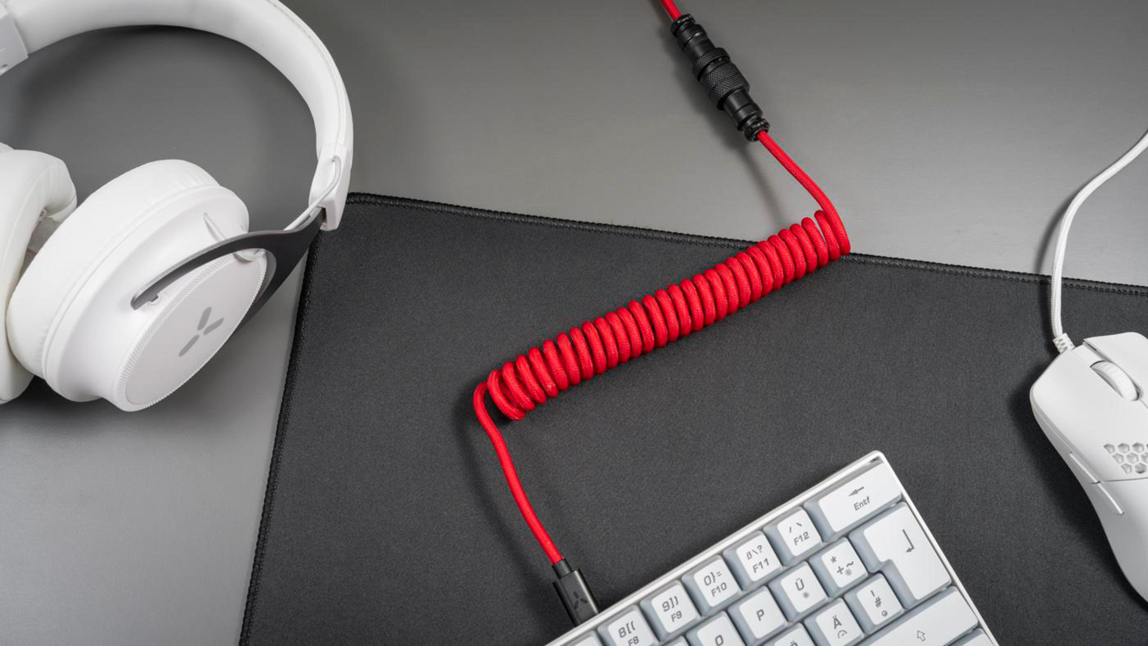 Kopfhörer, Tastatur und Maus auf einem Schreibtisch. Rotes Spiralkabel verbindet die Tastatur mit einem Gerät.