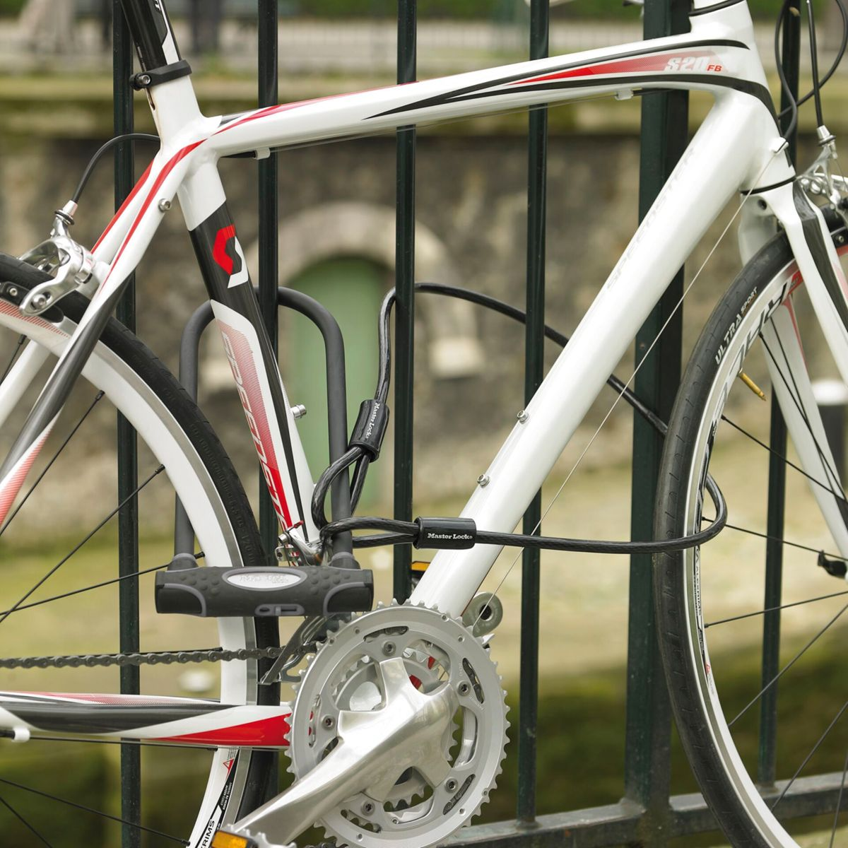 Ein weißes und rotes Fahrrad, das mit einem U-Schloss und Kabel an einem schwarzen Metallzaun befestigt ist.
