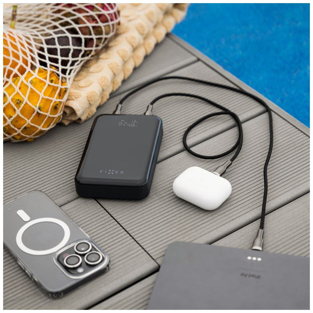 Eine schwarze Powerbank, weiße Ohrhörer und ein Telefon, die auf einer Pooloberfläche mit Früchten geladen werden.
