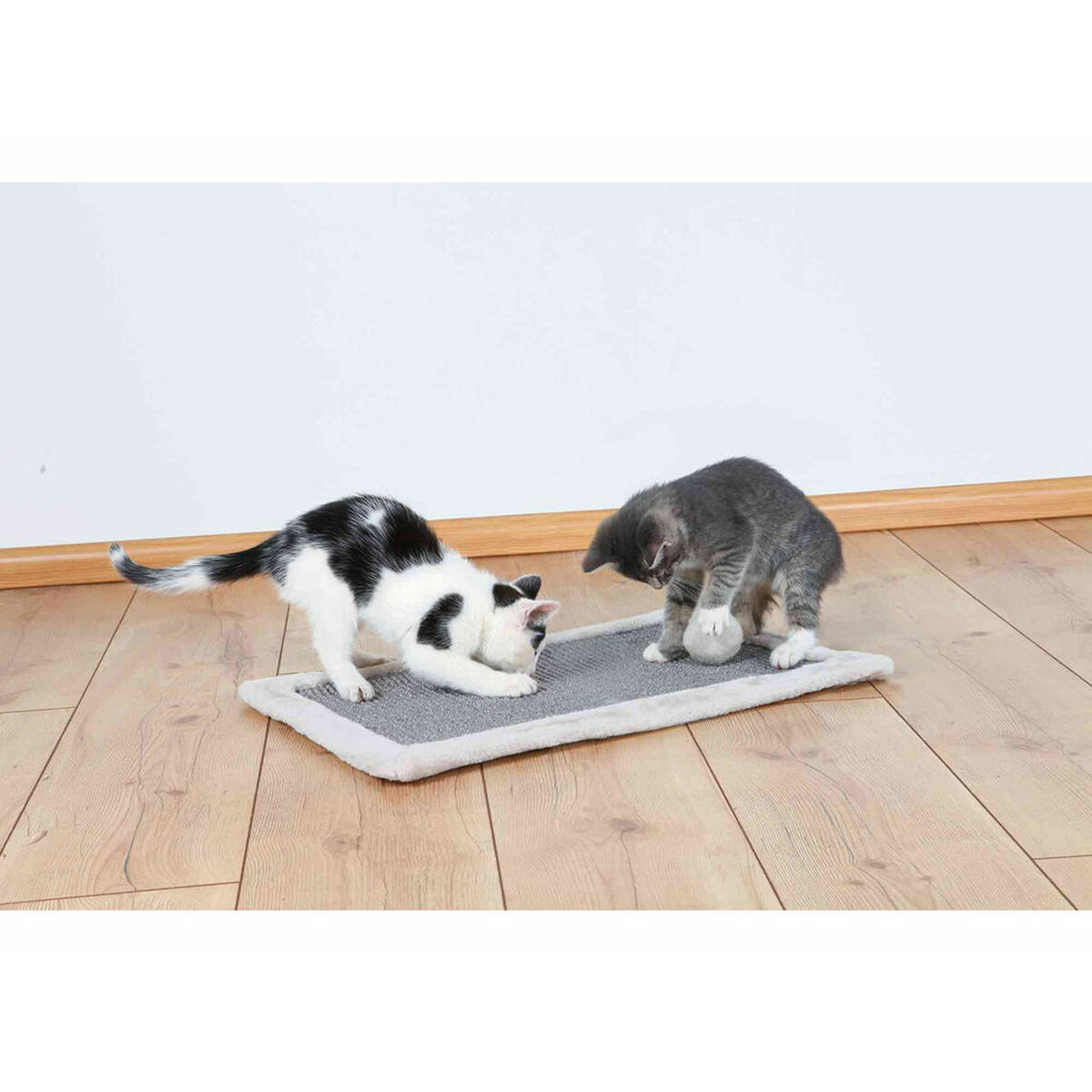 Dos gatitos jugando en una estera para rascar gris sobre un suelo de madera. Uno es blanco y negro, el otro gris.