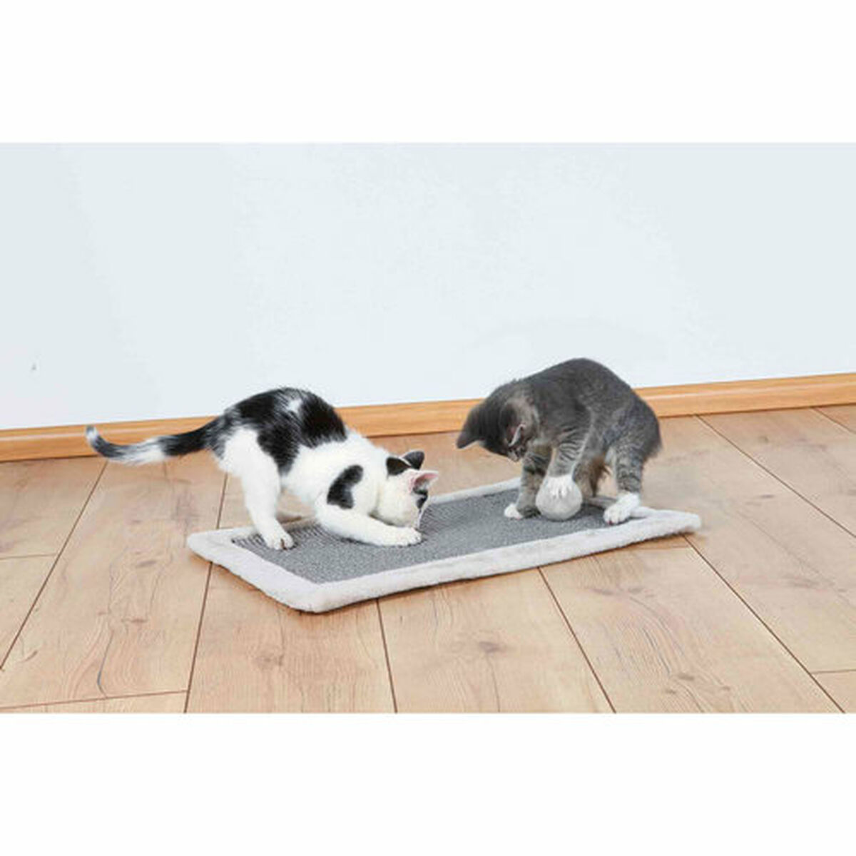 Dos gatos juegan en una alfombra gris sobre un suelo de madera. Uno es blanco y negro.