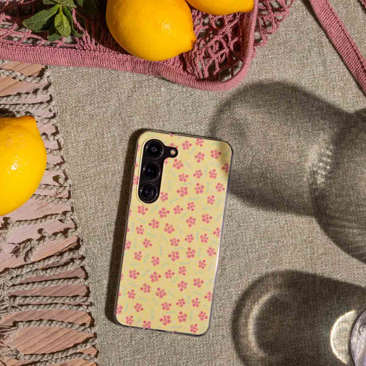 Gelbe Handyhülle mit rosa Blumenmuster, Zitronen und eine rosa Tasche auf einer Oberfläche.