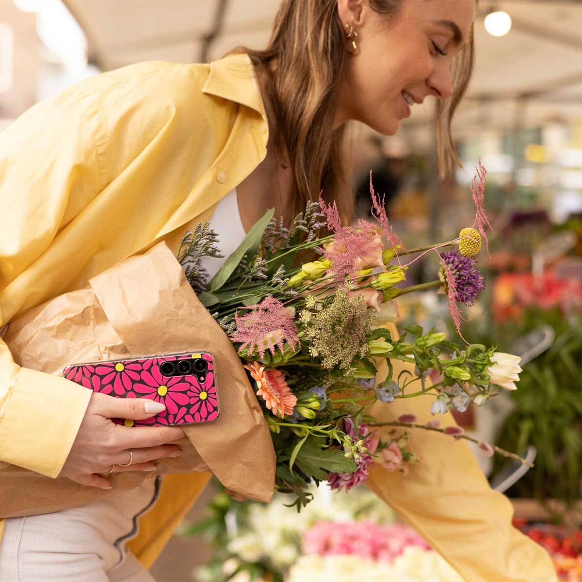 Frau in gelbem Hemd hält einen Blumenstrauß und eine Handytasche mit rosa Blumen auf einem Blumenmarkt.