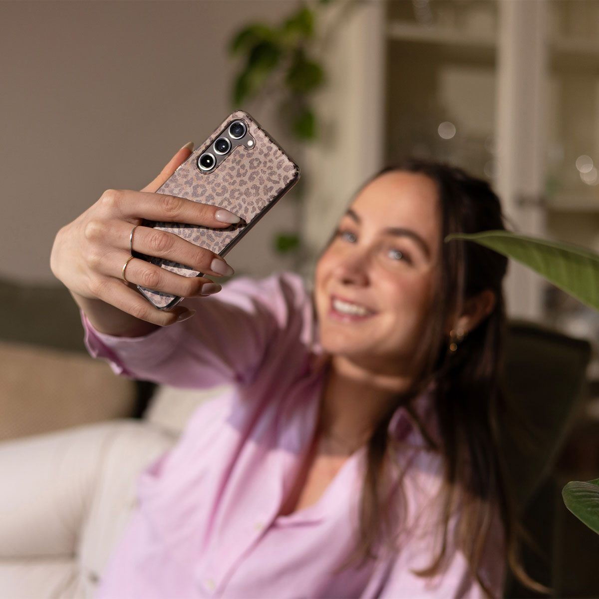 Frau macht Selfie mit Leopardenmuster-Handyhülle, lächelnd. Pflanzen im Hintergrund.