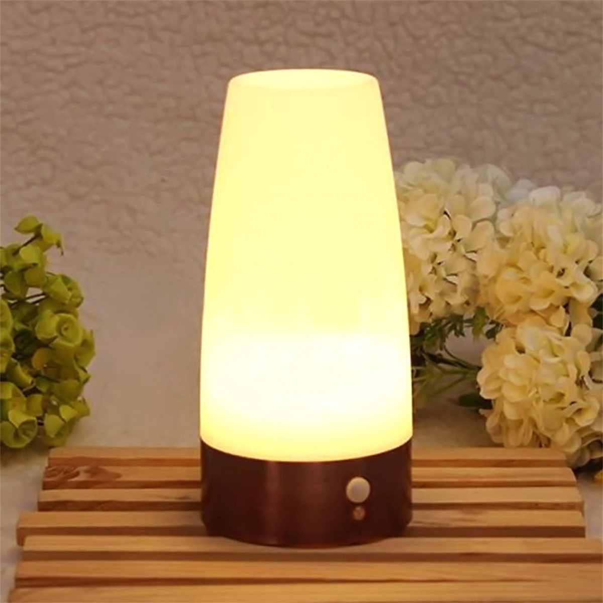 Eine leuchtende weiße Lampe mit braunem Sockel, die auf einem Holzständer steht, mit Blumen im Hintergrund.