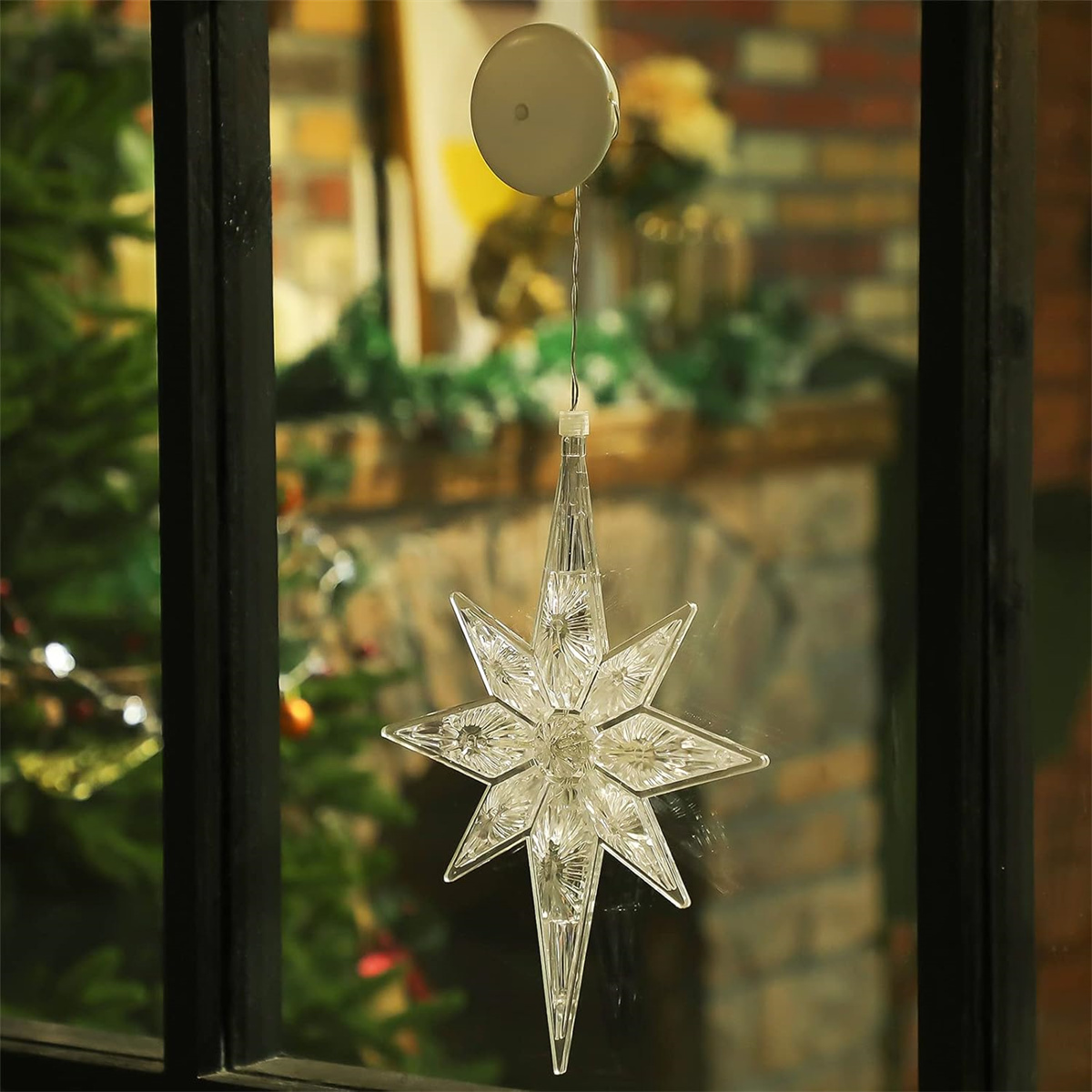 Ein beleuchteter, klarer Plastikstern hängt vor einem Fenster, mit einem Weihnachtsbaum sichtbar.