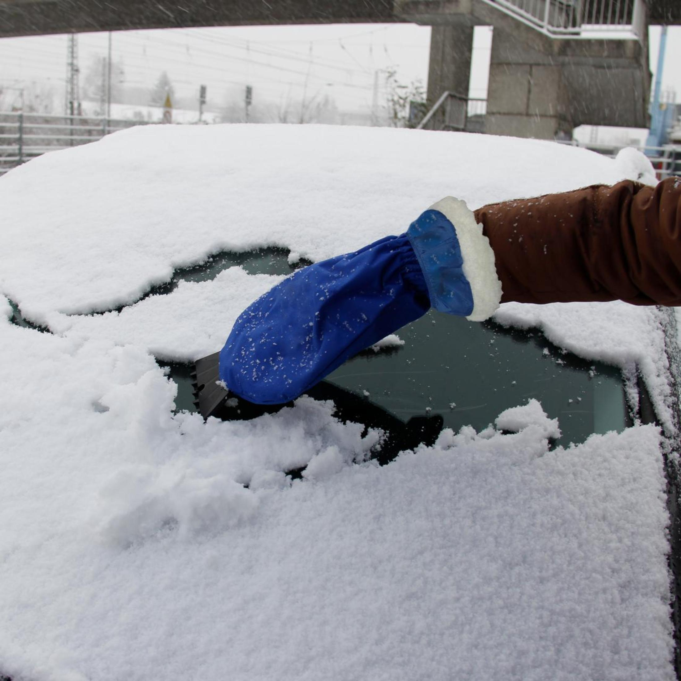 Eine Hand kratzt Schnee mit einem blauen Handschuh und einem Schaber von einer Autoscheibe ab.