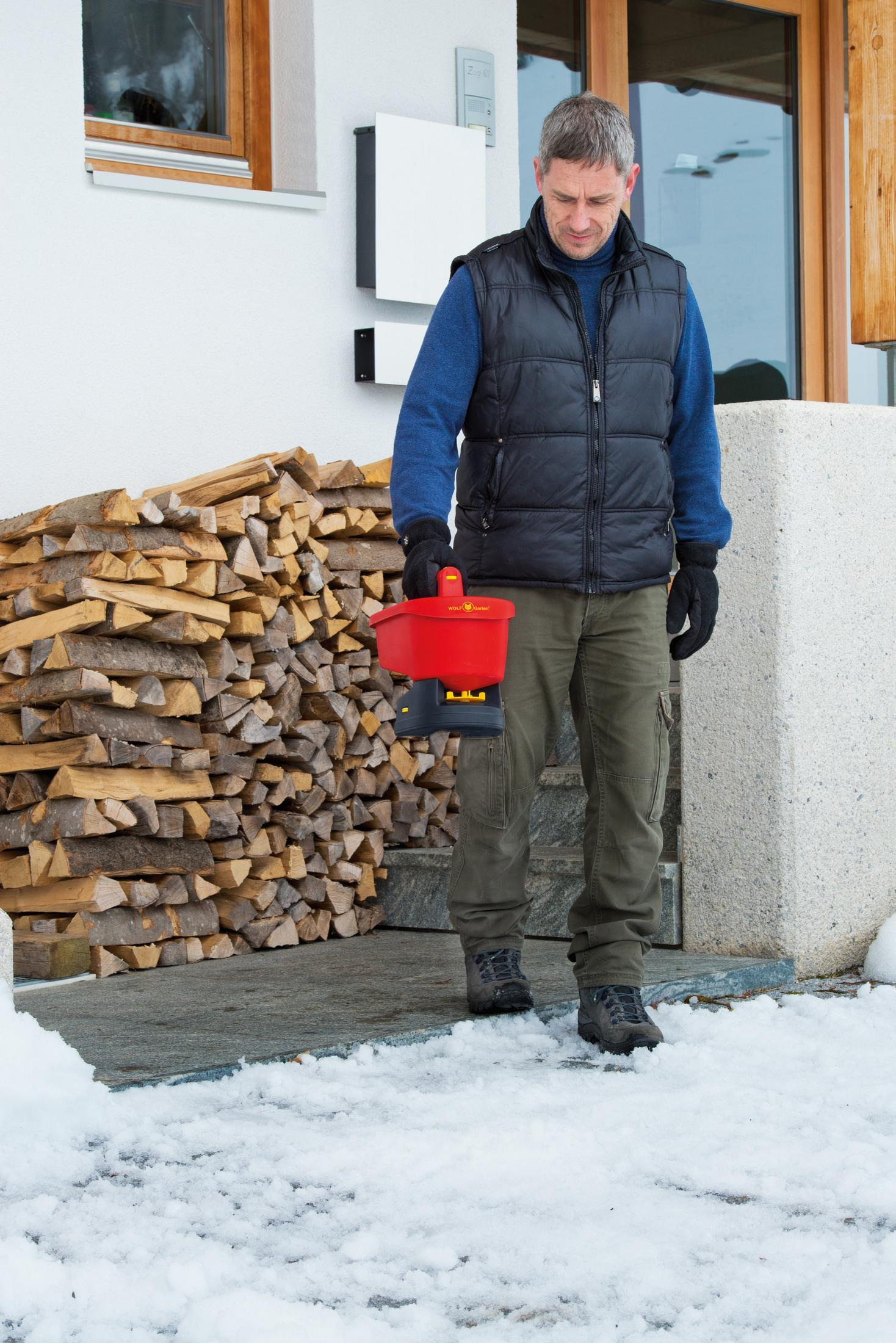 Ein Mann geht auf einem verschneiten Weg und hält einen roten Behälter. Im Hintergrund Holz.