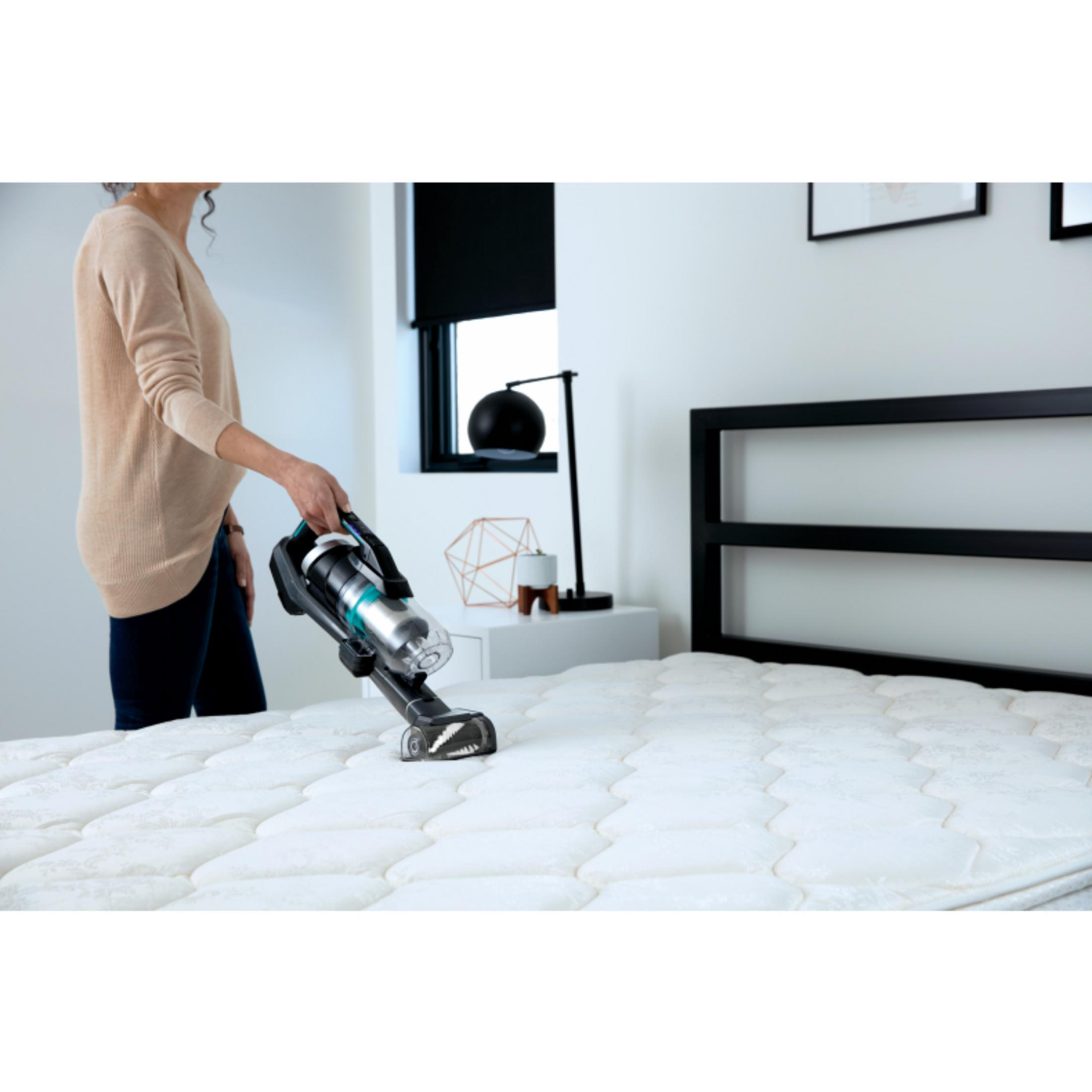 Donna che usa un aspirapolvere su un letto bianco. Comodino, finestra e tenda nera.