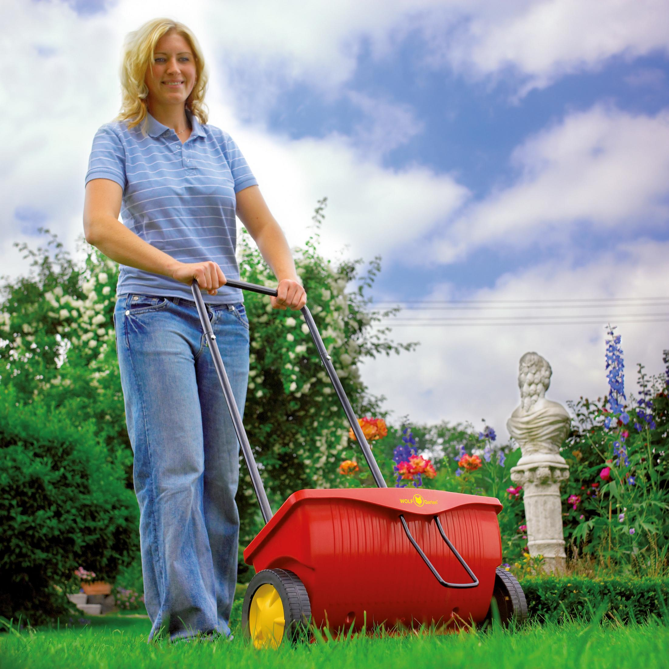 Frau schiebt einen roten Rasenstreuer auf grünem Gras. Blumen und blauer Himmel im Hintergrund.
