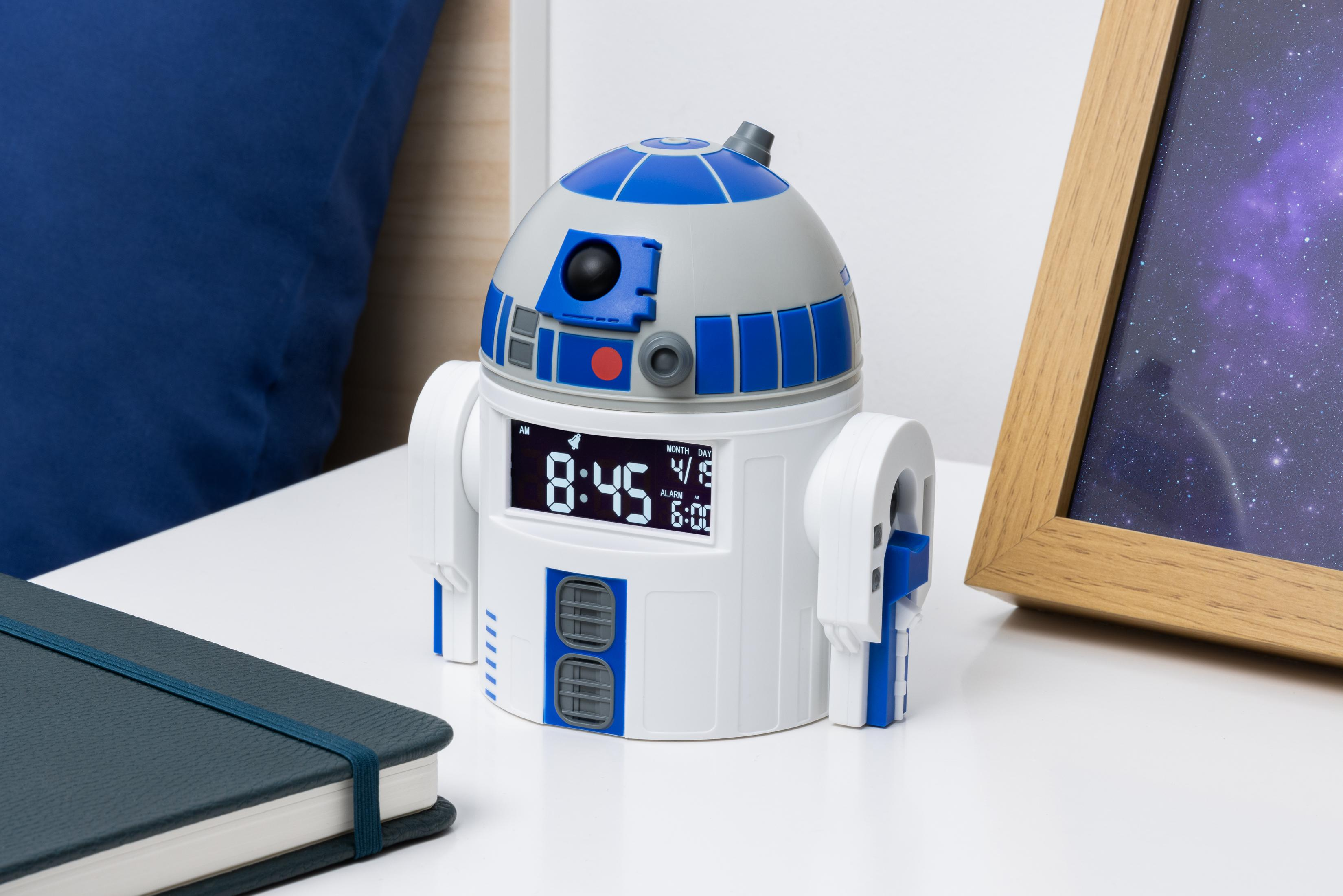Budzik R2-D2 na stoliku nocnym. Zegar jest biało-niebieski, obok notatnika i ramki na zdjęcia.