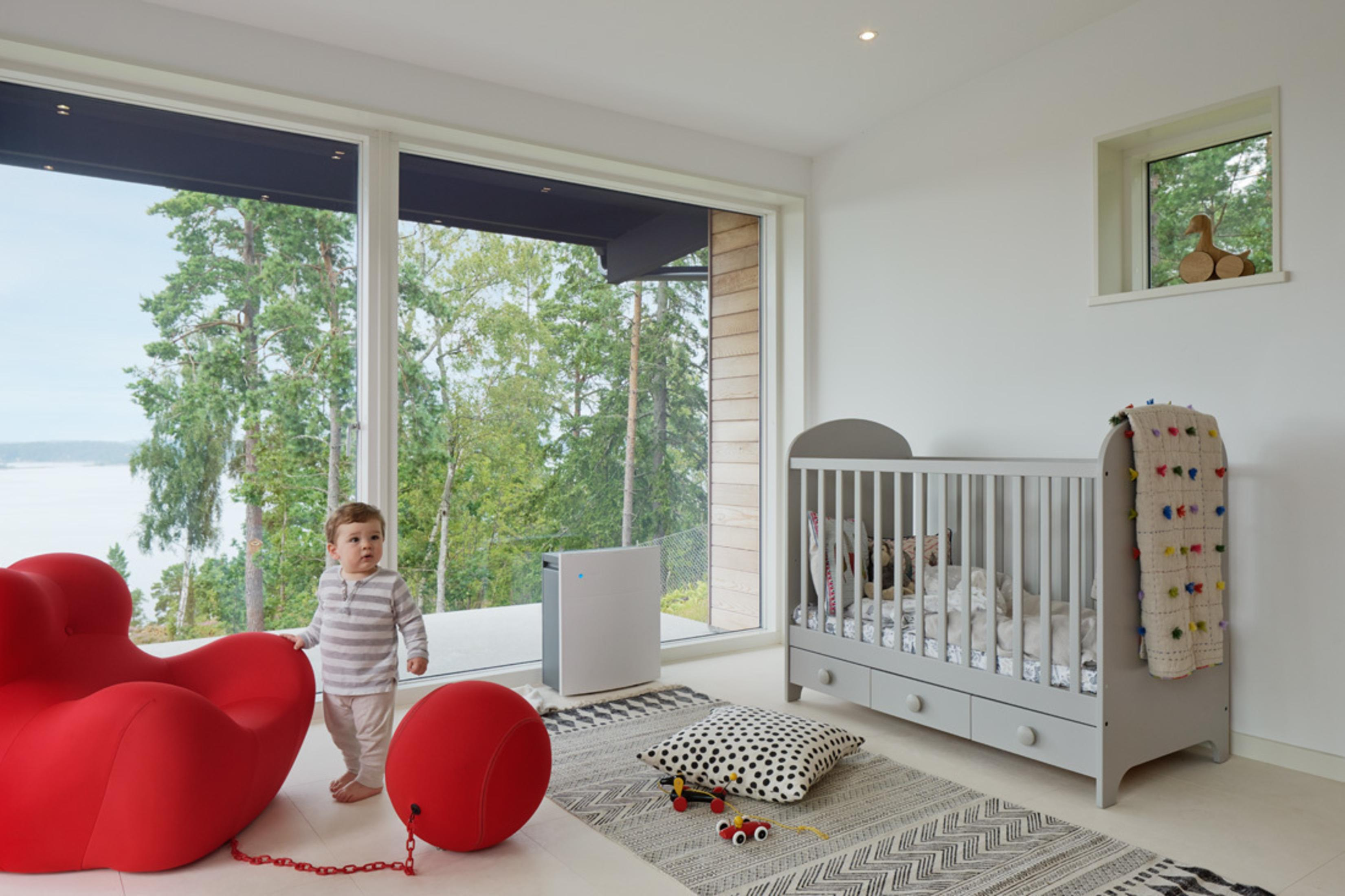 Ein Kind in einem Zimmer mit Kinderbett, rotem Stuhl, großem Fenster und Landschaft.