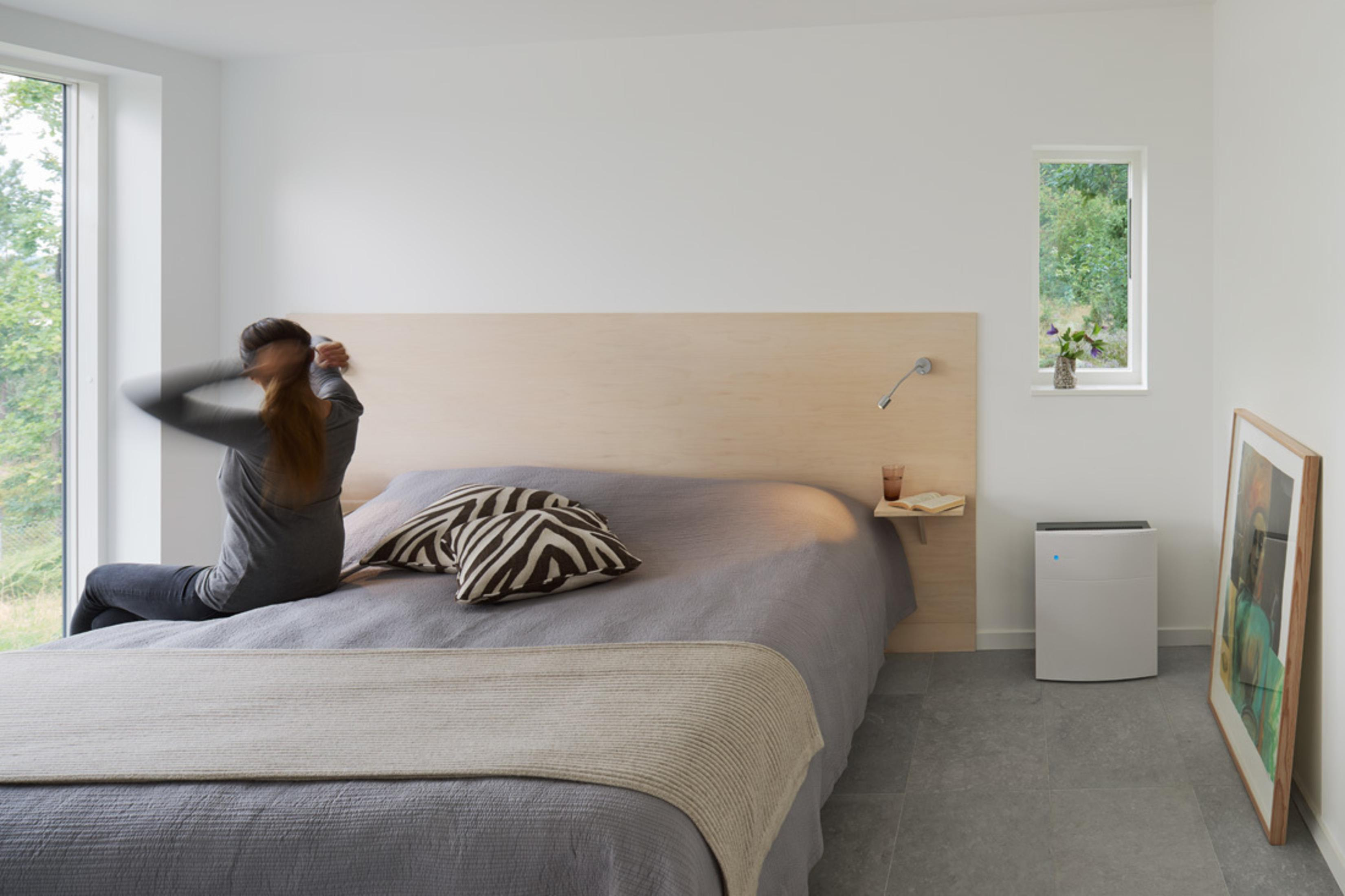 Frau im Schlafzimmer, zum Fenster greifend, mit Bett und Luftreiniger.
