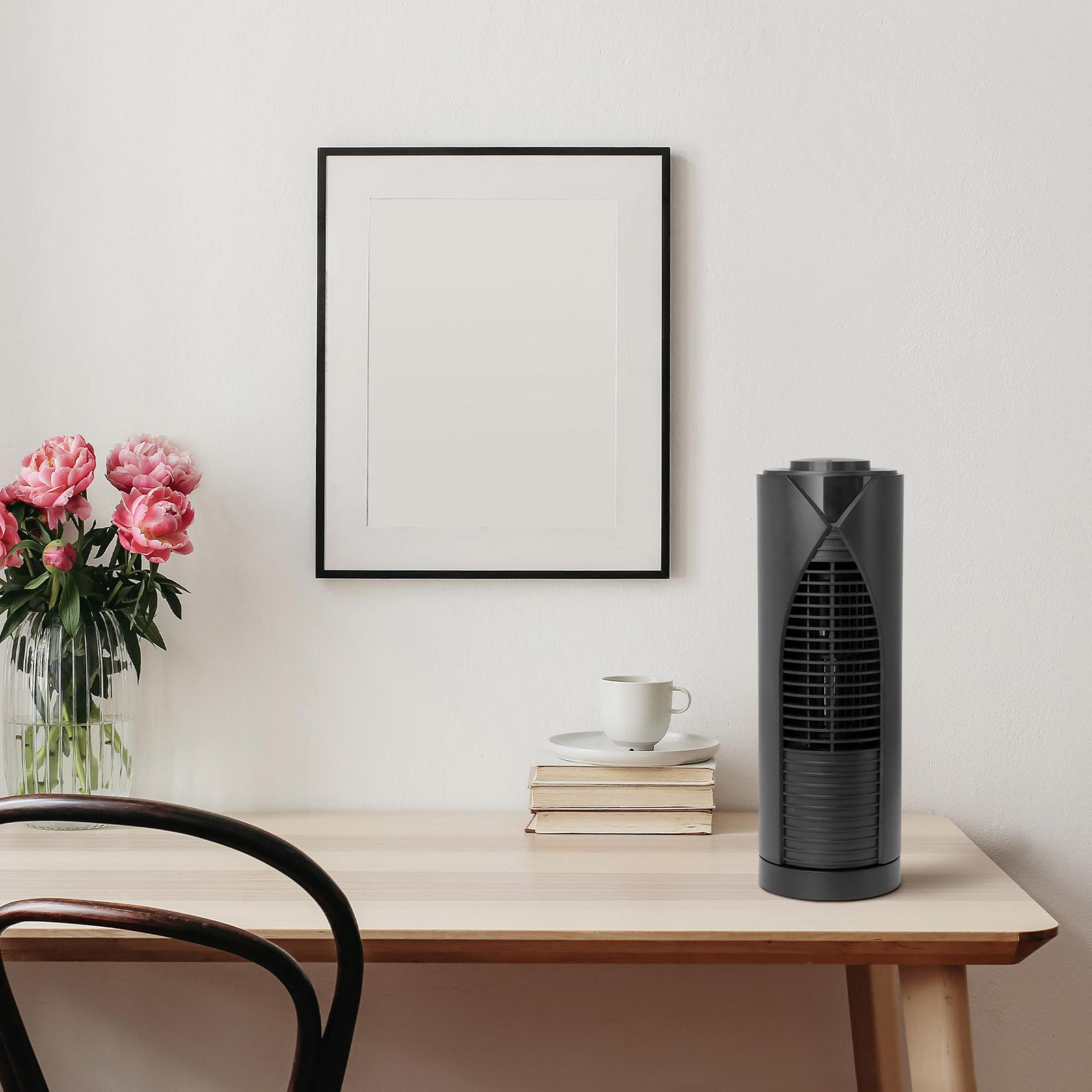 Ein schwarzer Turmventilator steht auf einem Holztisch. Dahinter ein gerahmtes Bild und eine Vase mit rosa Blumen.