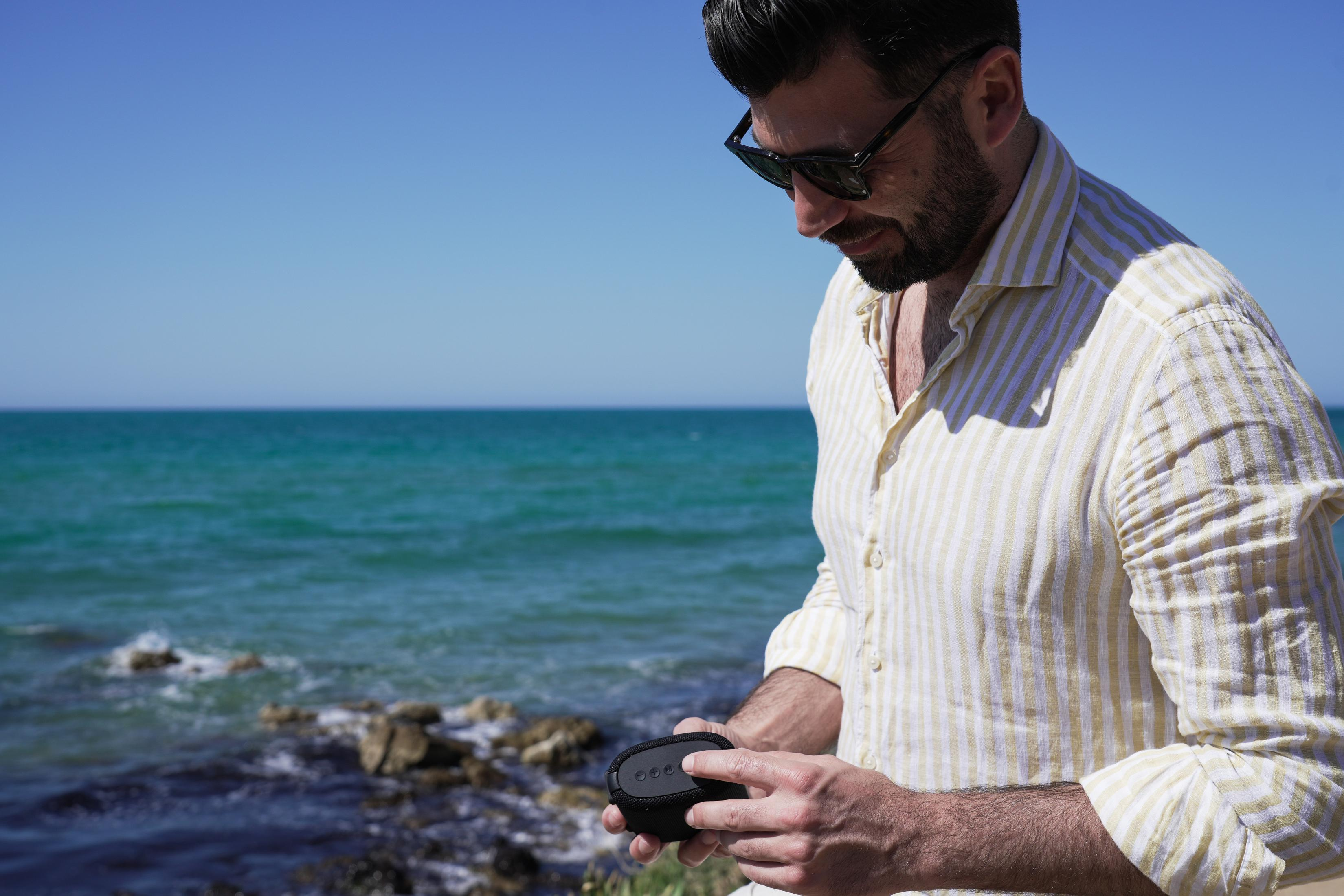 Mann mit Sonnenbrille am Meer mit Lautsprecher in der Hand. Das Meer ist türkis und der Himmel blau.