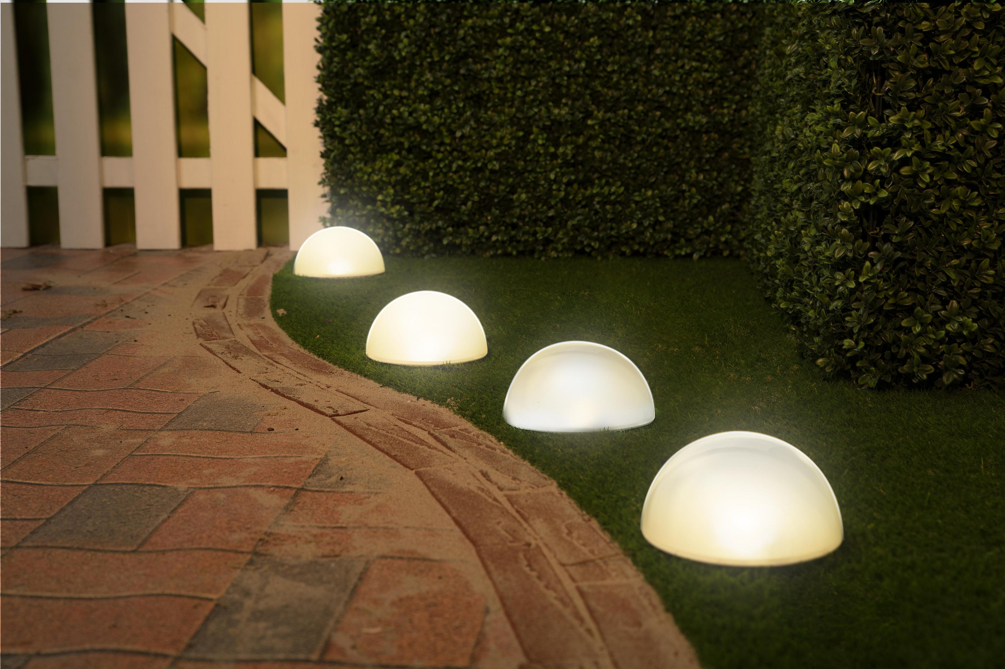 Quatre lumières en forme de demi-sphère illuminent une pelouse verte près d'une clôture blanche et d'un chemin de briques.