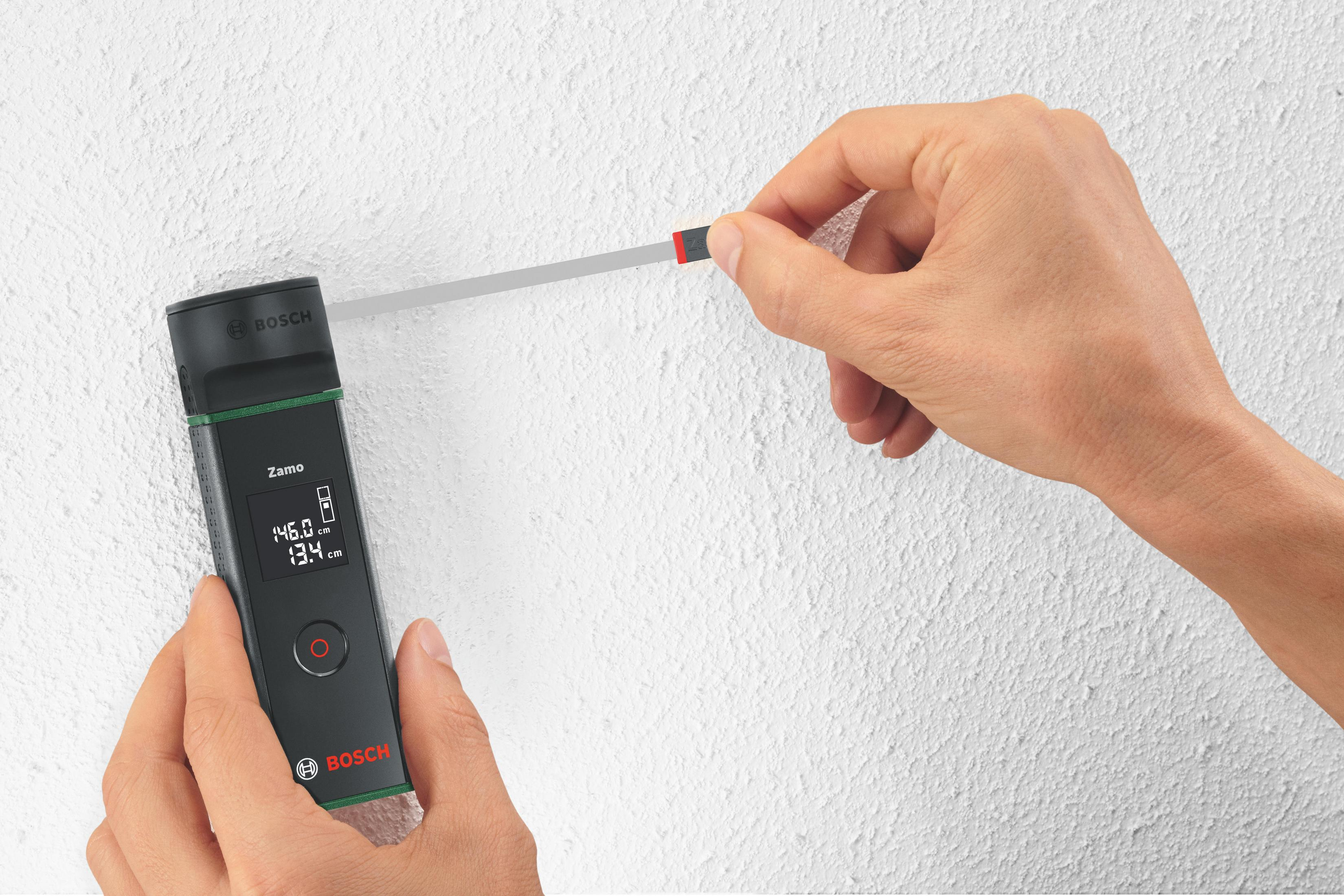 Bir kişi, beyaz bir duvara karşı siyah ve yeşil bir Bosch Zamo lazer mesafe ölçer tutuyor.