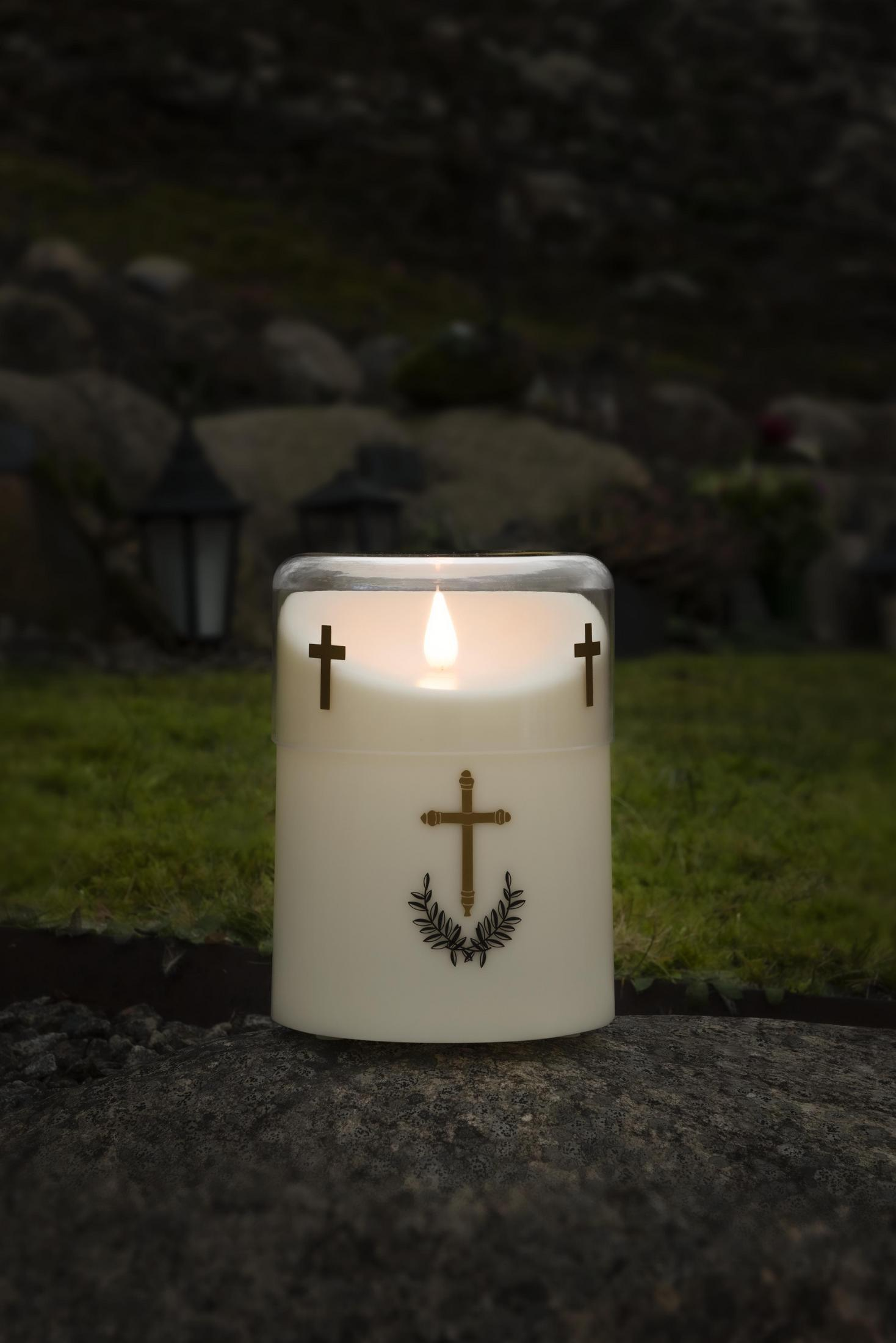 Eine brennende Kerze mit Kreuzdesign auf der Vorderseite. Sie steht auf einer Steinfläche vor einem Friedhof.