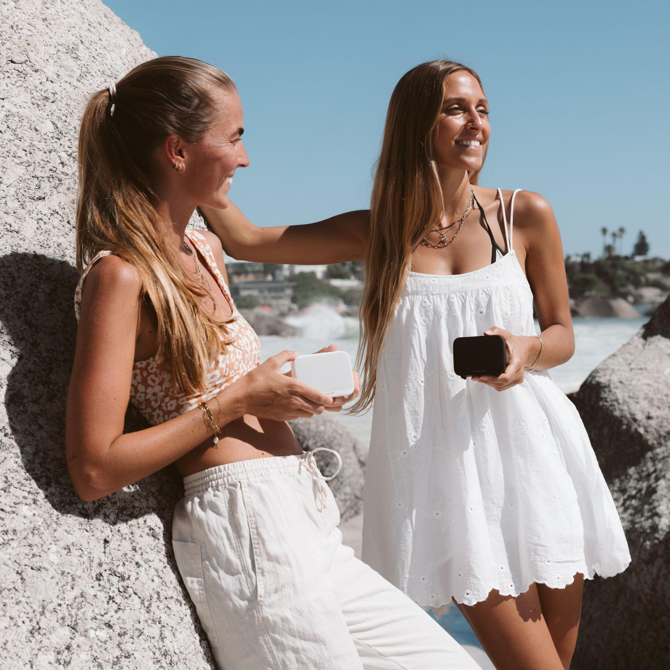 Zwei Frauen am Strand, eine hält einen weißen Lautsprecher, die andere einen schwarzen. Der Hintergrund ist ein sonniger Strand.