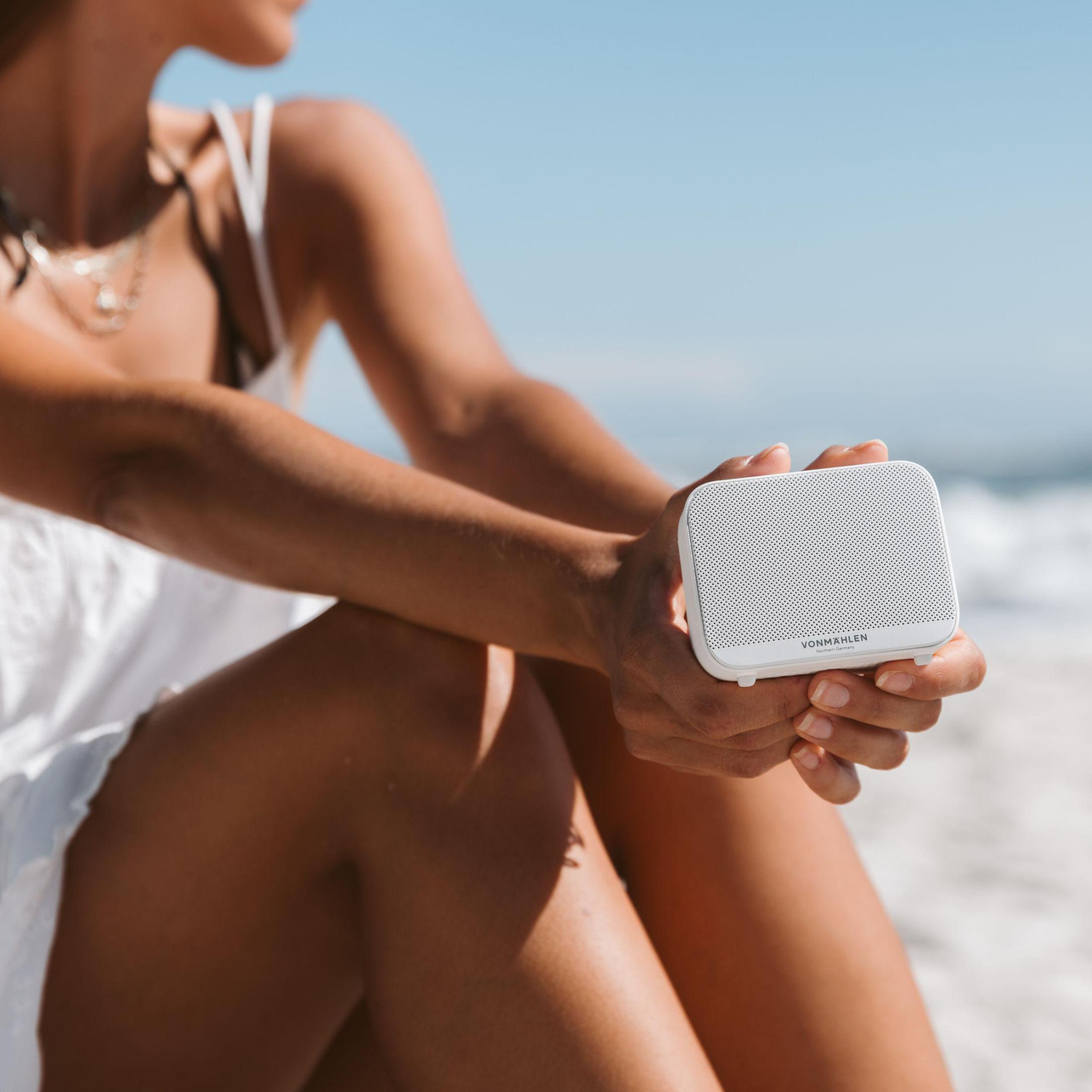 Eine Frau hält einen kleinen weißen Lautsprecher am Strand. Strand und Meer sind sichtbar.