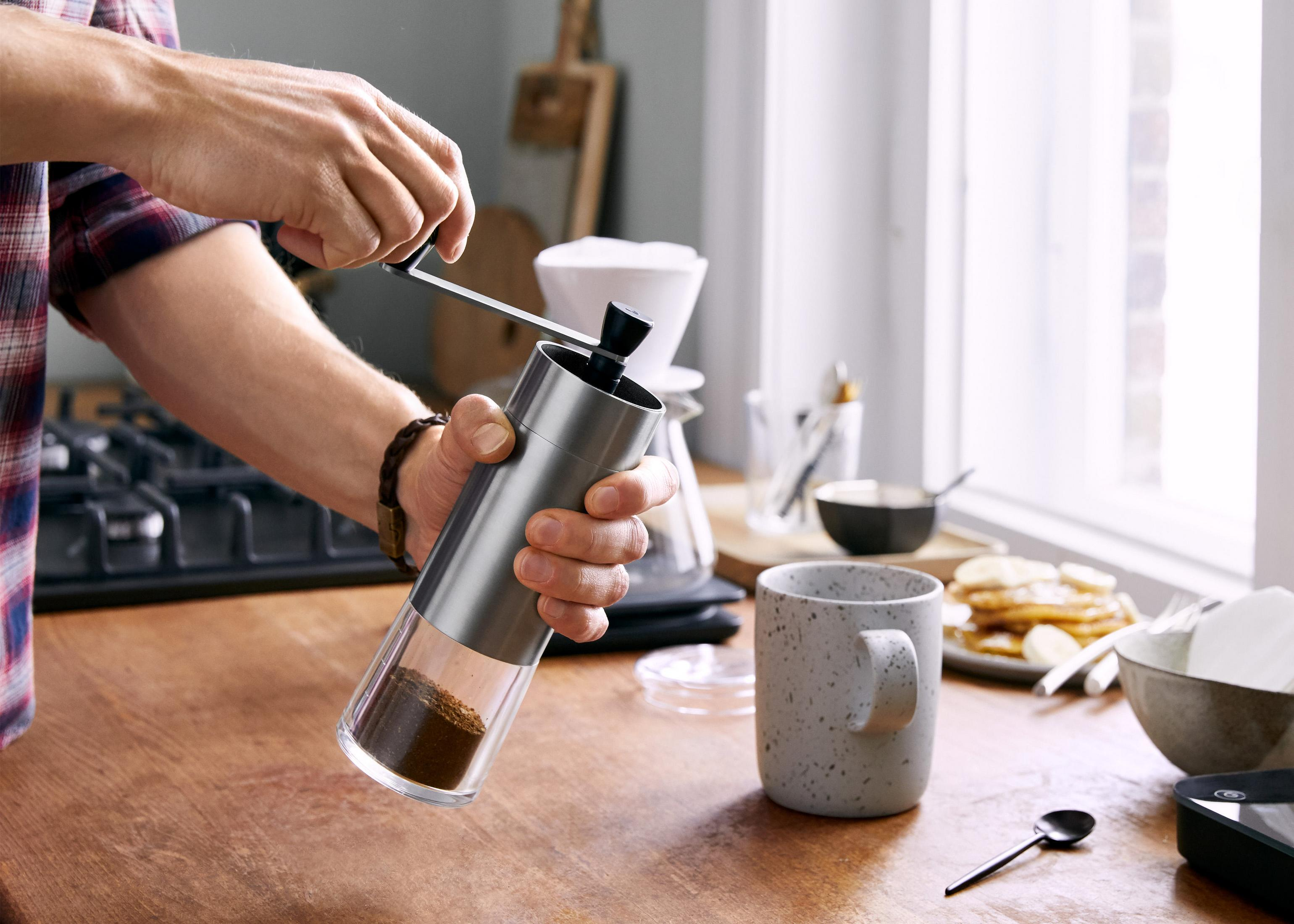 Person mahlt Kaffeebohnen mit Handmühle, Tasse und anderen Gegenständen.