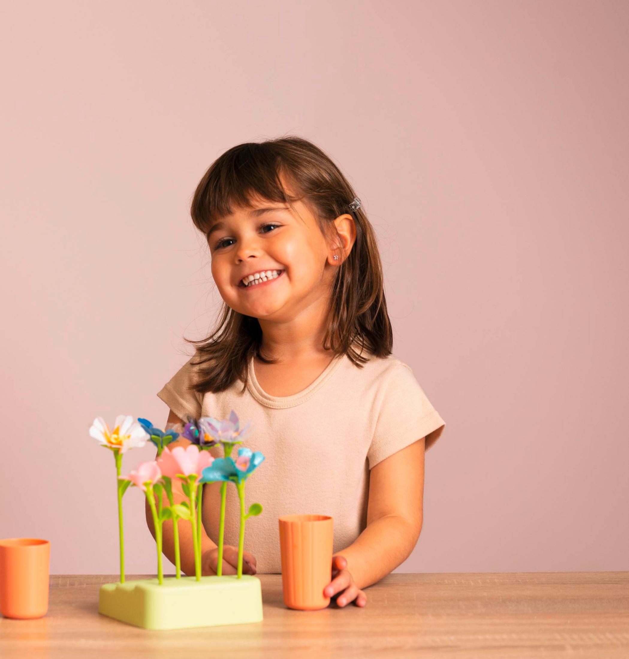 Ein lächelndes Mädchen mit Blumen und orangefarbenen Bechern auf einem Holztisch, vor pinkfarbenem Hintergrund.