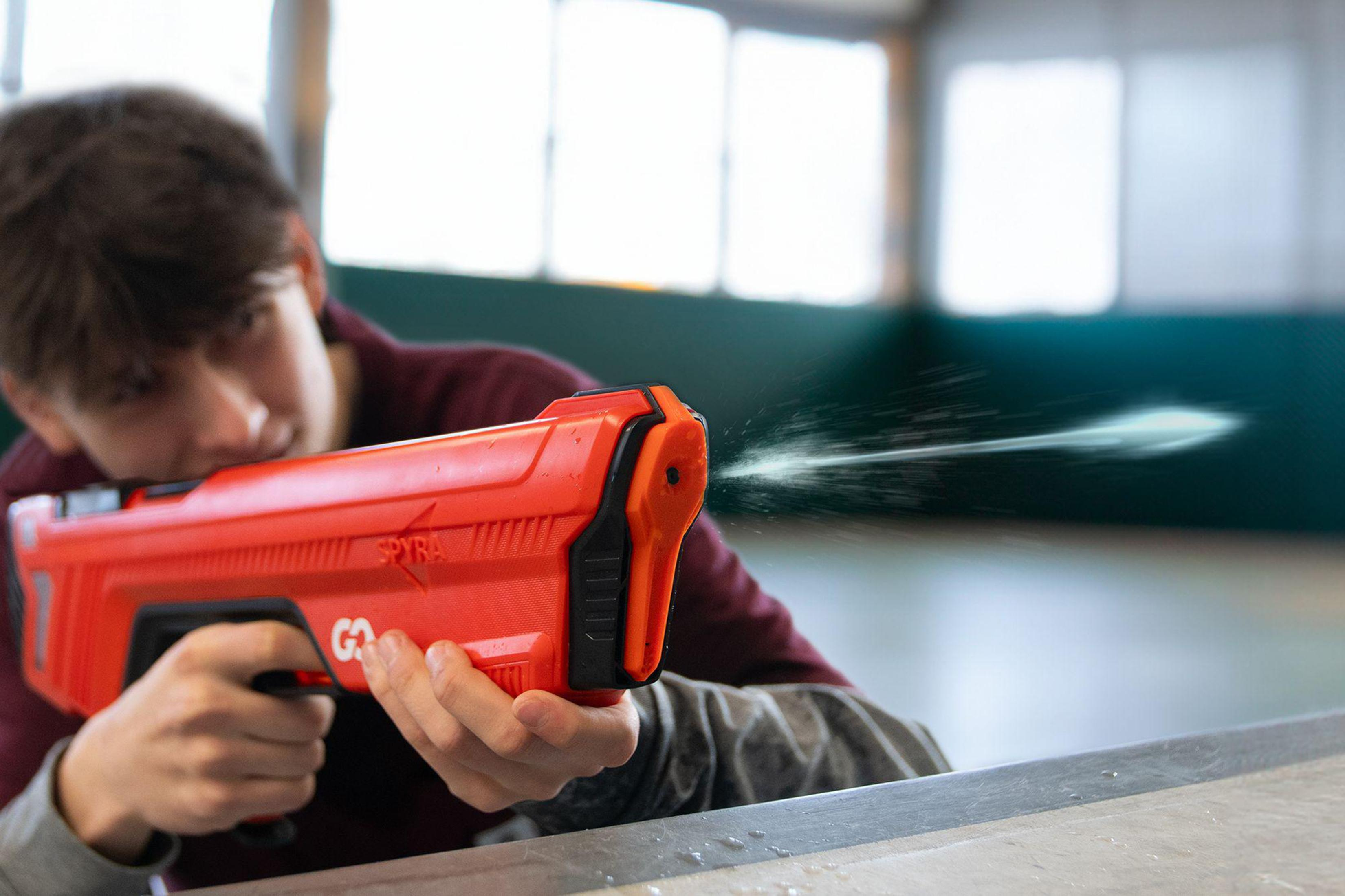 Junge schießt Wasser aus einer rot-schwarzen Wasserpistole in einer Turnhalle.