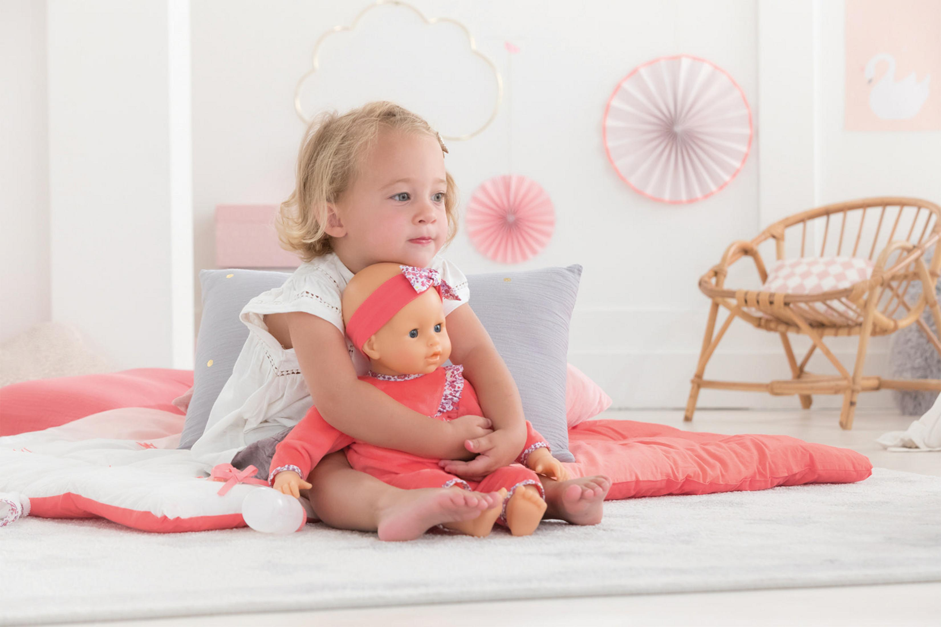Blondes Mädchen umarmt eine Puppe in einem Zimmer mit rosa und weißer Einrichtung.