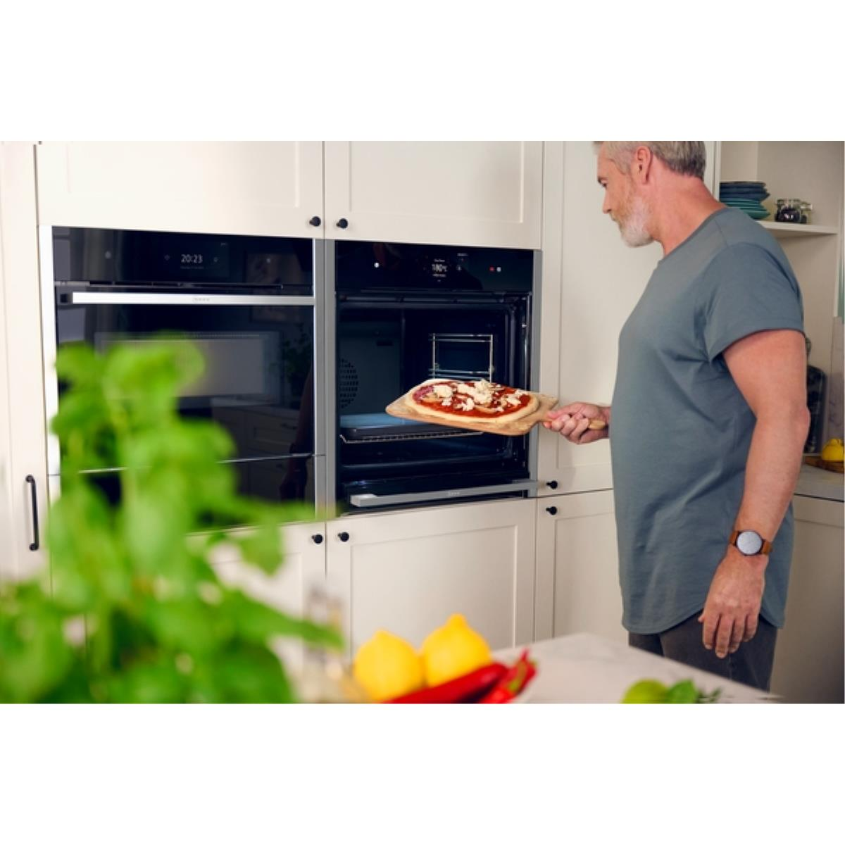 Mann, der Pizza in den Ofen legt. Küche und Essen im Fokus. Grüne Pflanze im Vordergrund.
