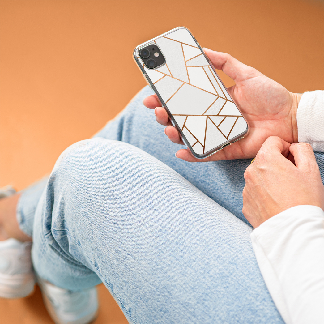 Une personne tenant un téléphone avec une coque géométrique blanche et dorée, assise sur un jean.
