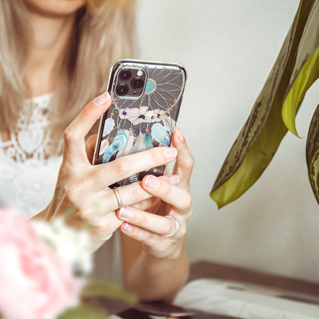 Een vrouw houdt een telefoon vast met een dromenvanger hoesje en een plant op de achtergrond.