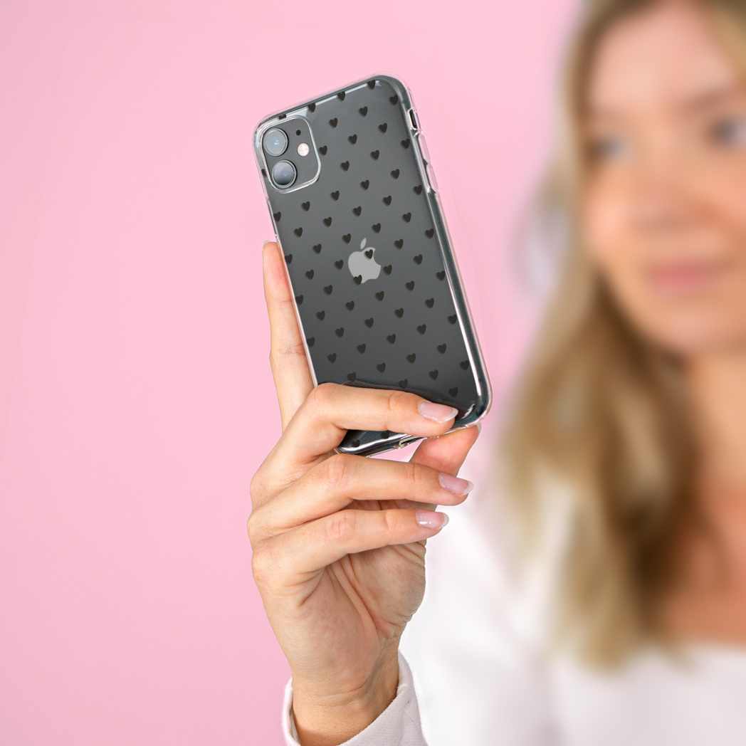 Una donna tiene in mano un telefono con una custodia coperta da piccoli cuori neri. Lo sfondo è rosa.