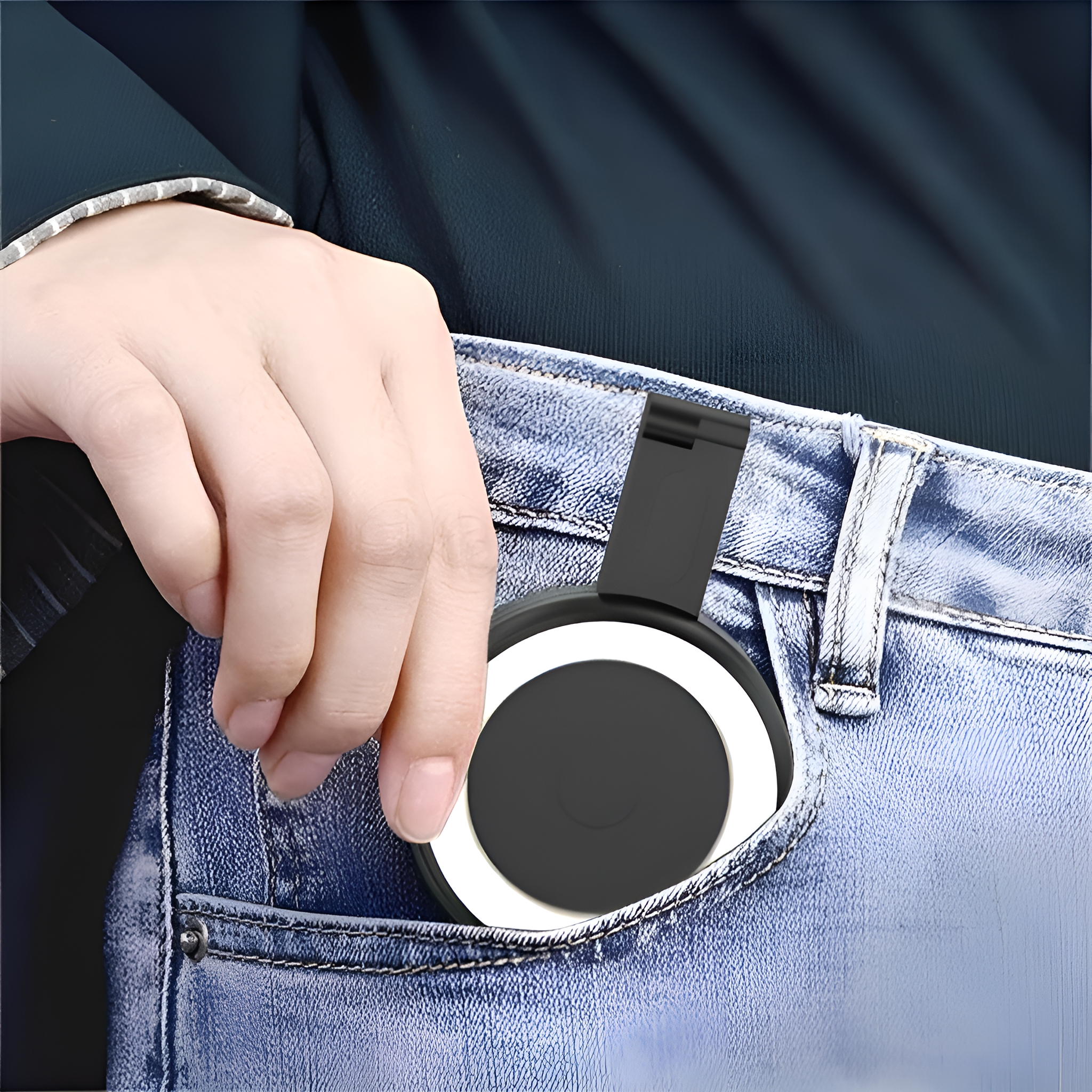 Hand hält einen schwarzen, zusammenklappbaren Telefonständer in einer Jeans-Tasche. Der Ständer ist rund und schwarz-weiß.