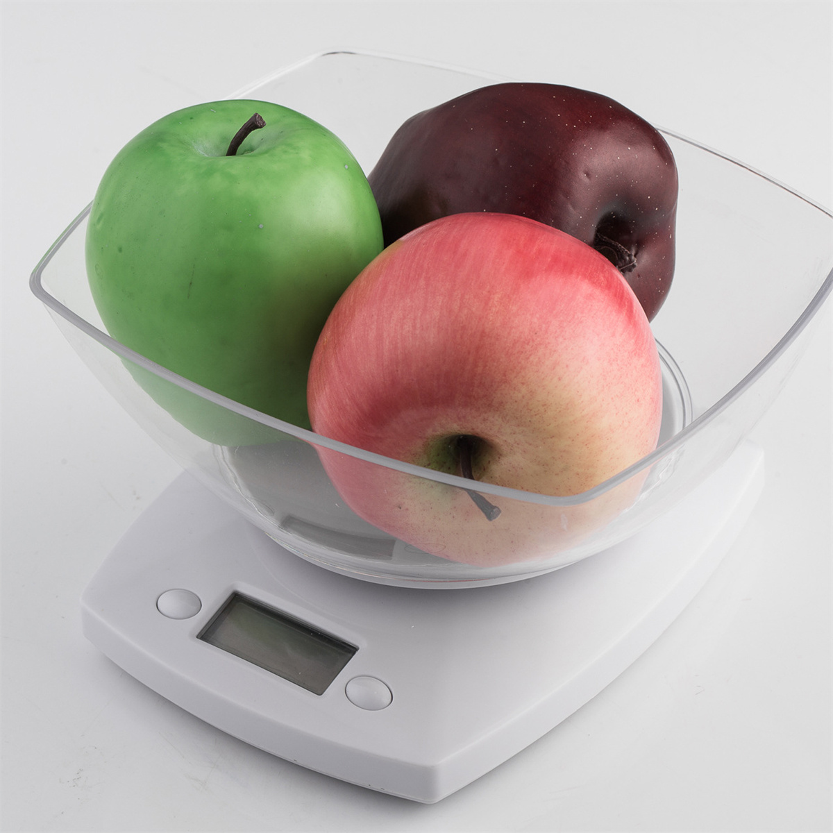 Drei Äpfel in einer durchsichtigen quadratischen Schüssel auf einer weißen Waage. Ein grüner, ein roter, ein dunkelroter Apfel.
