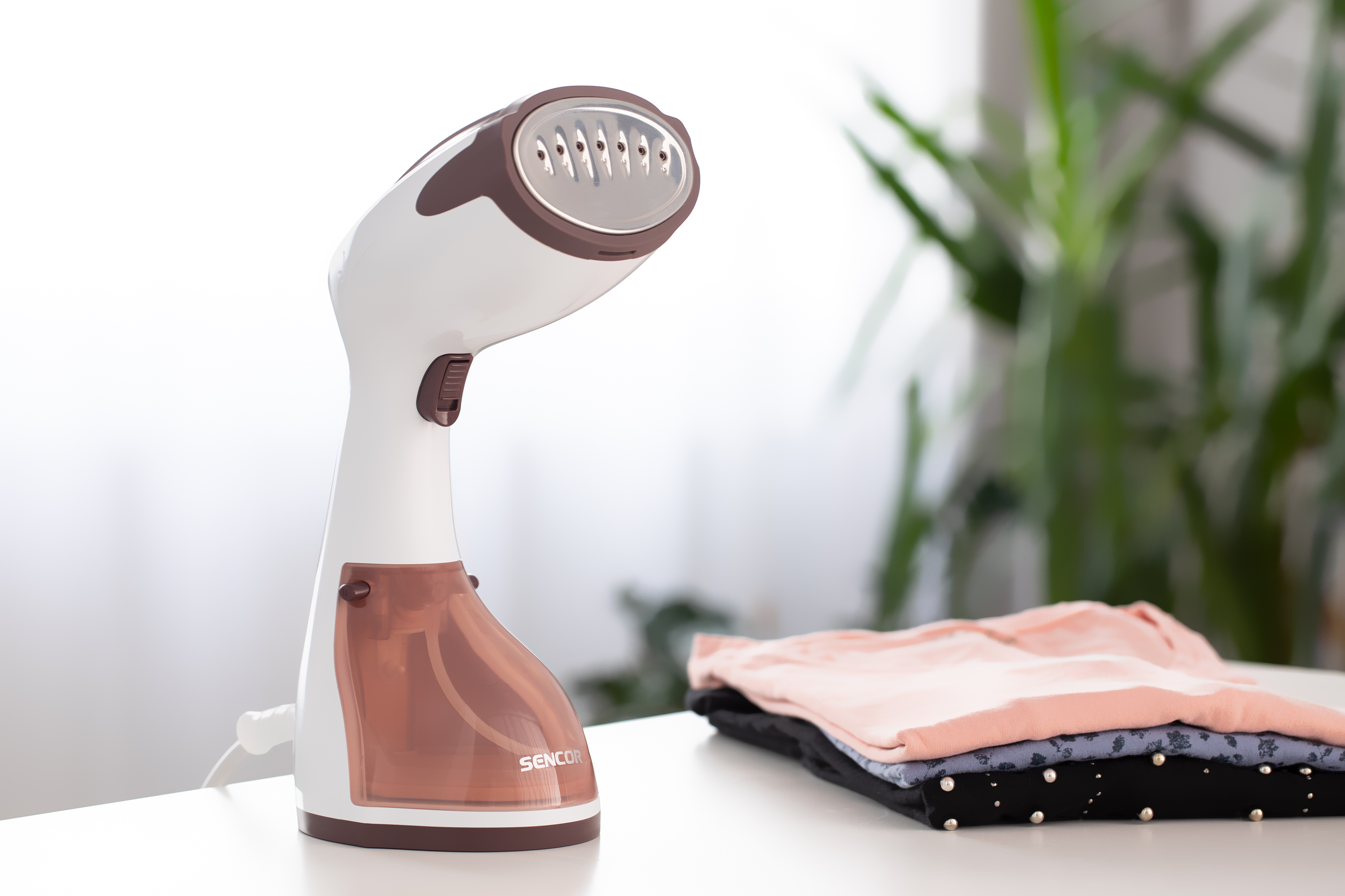 Un vaporizzatore per indumenti bianco e marrone accanto a una pila di vestiti su un tavolo bianco.
