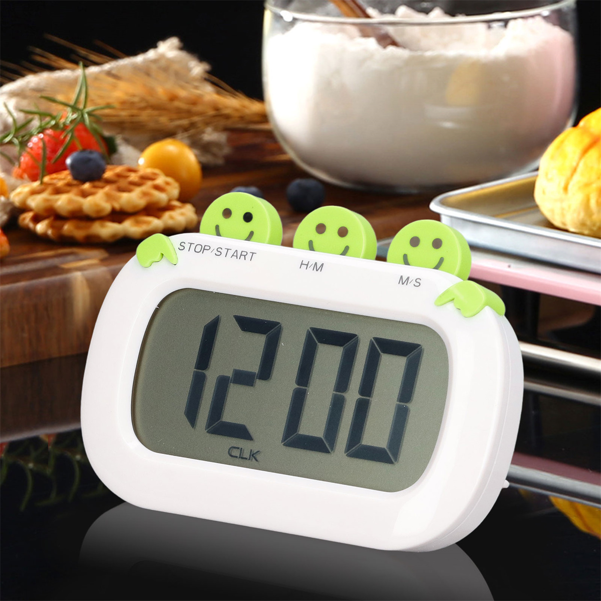 Weißer Küchentimer mit grünen Smiley-Knöpfen. Zeigt 12:00 an. Backwaren im Hintergrund.