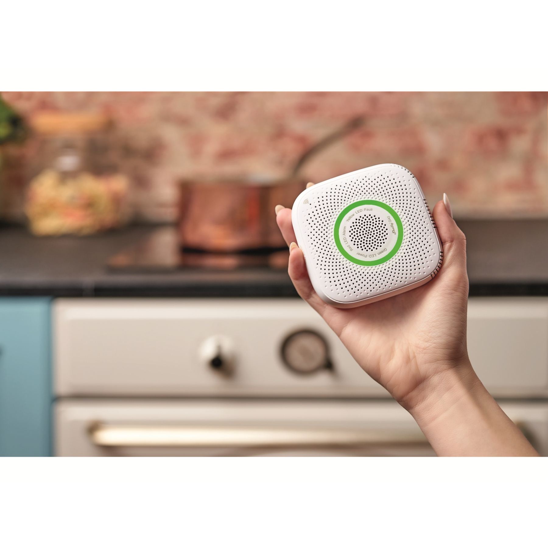 Dłoń trzymająca biały oczyszczacz powietrza z zielonymi akcentami, przed kuchnią z miedzianymi garnkami.