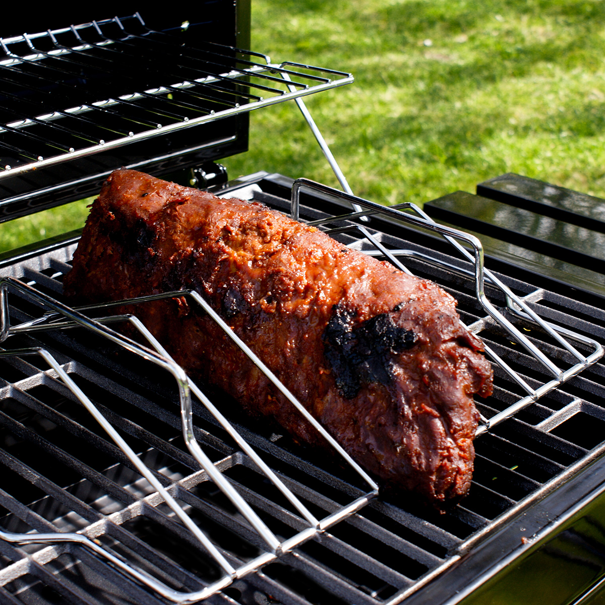A cooked, glazed meat loaf is on a grill. The grill is on a black frame, sitting on grass.