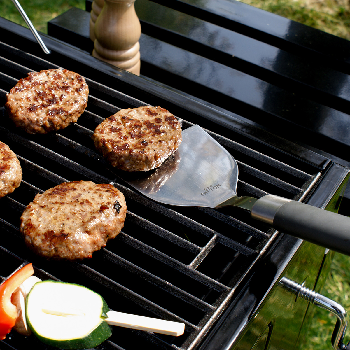 Burgers on a grill, a spatula is lifting one. Zucchini and peppers are beside.